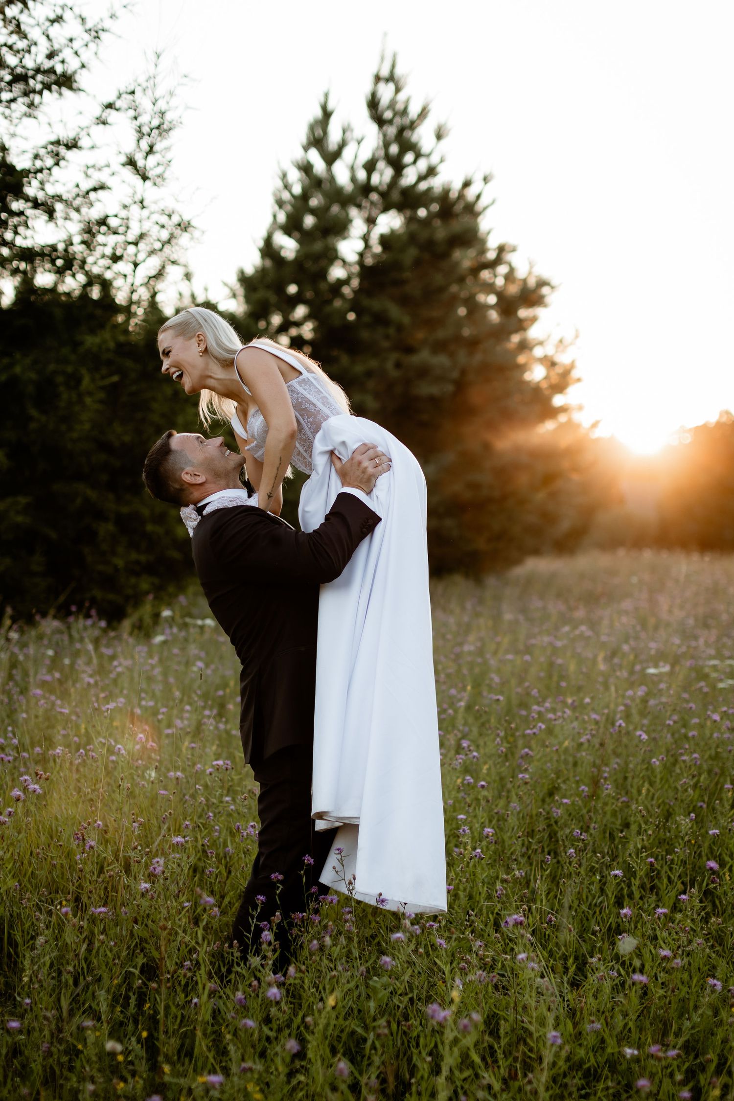 Romantic wedding lift pose in a sunlit meadow with evergreen trees in background.