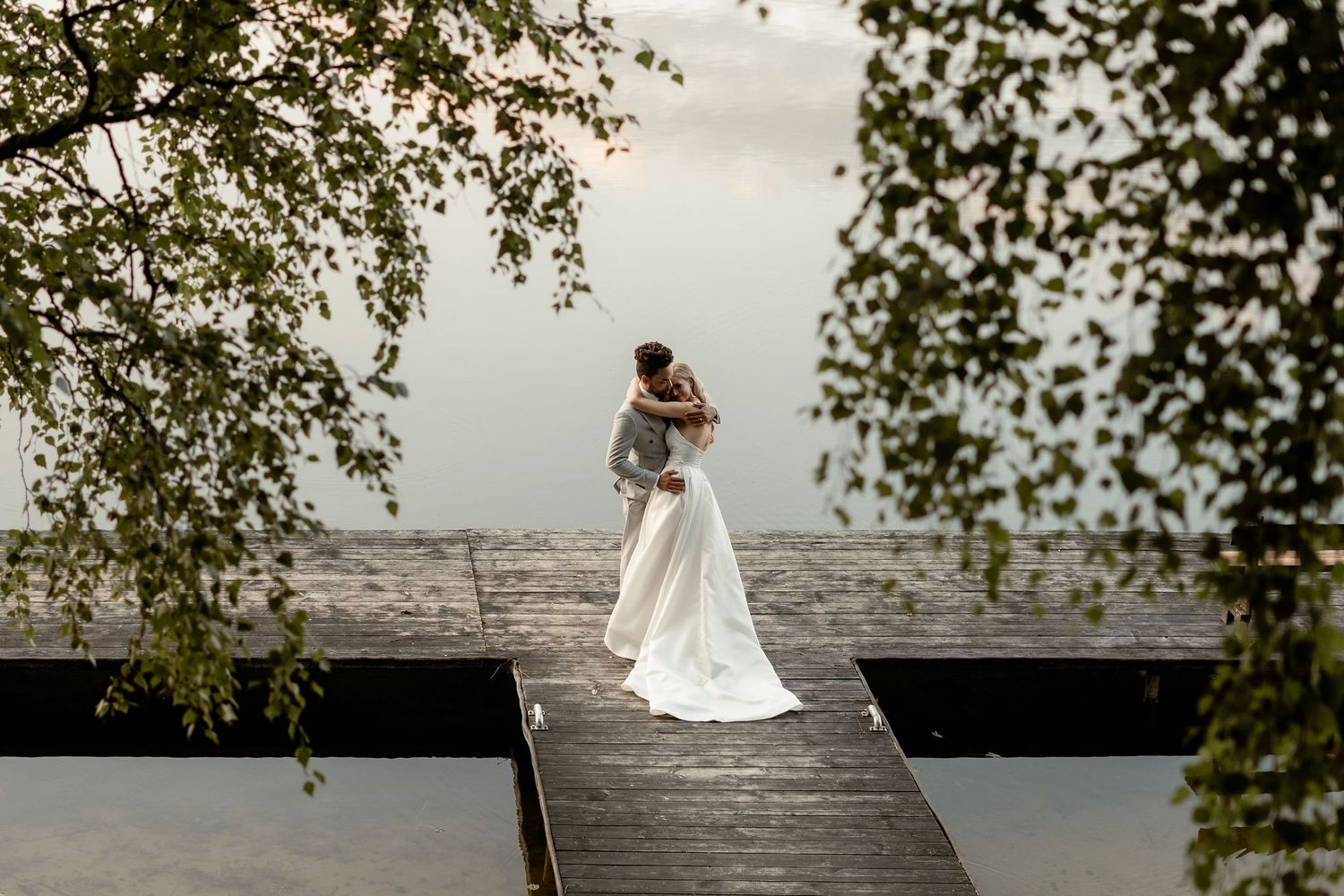 Romantic wedding couple embraces on wooden dock over misty lake with overhanging tree branches framing the scene.