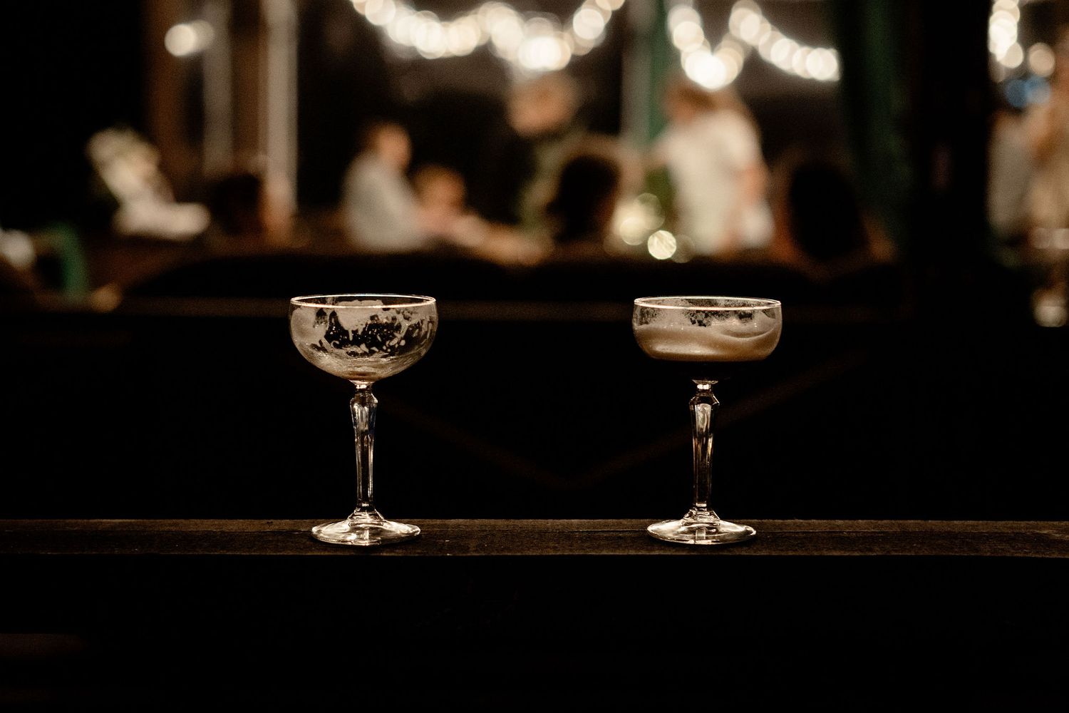 Two elegant cocktail glasses sit on a dark wooden bar with soft bokeh lights in the moody background.
