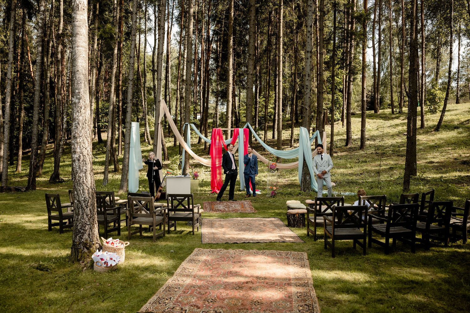 Outdoor wedding ceremony setup in a pine forest with blue fabric draping and wooden chairs along an aisle.