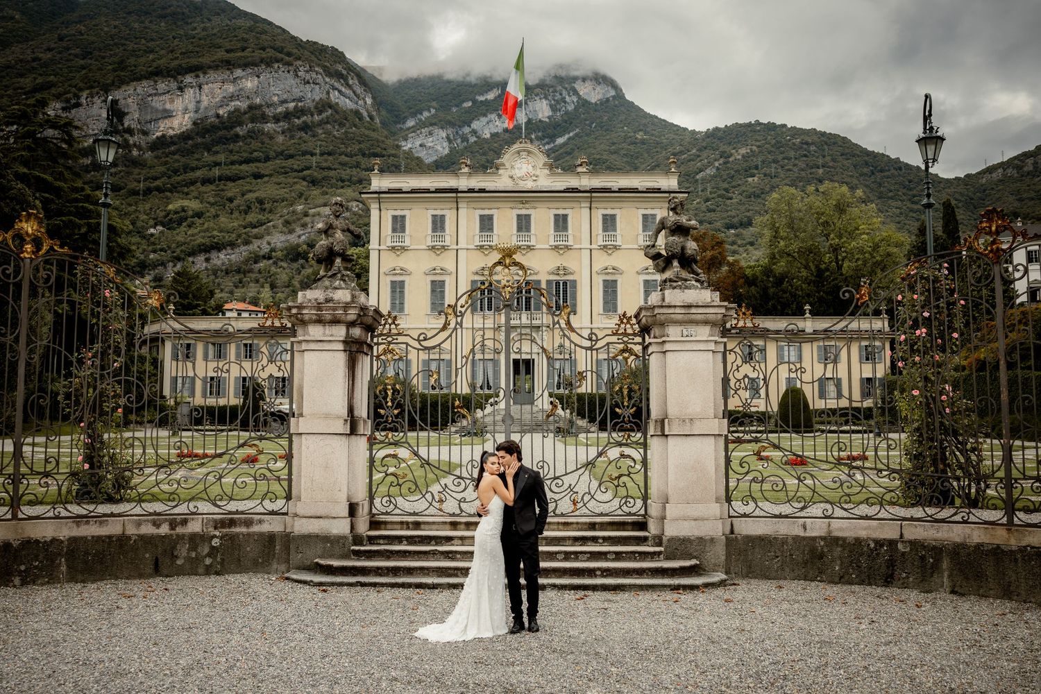 Wedding couple poses in front of grand historic villa with mountain backdrop and ornate gates.