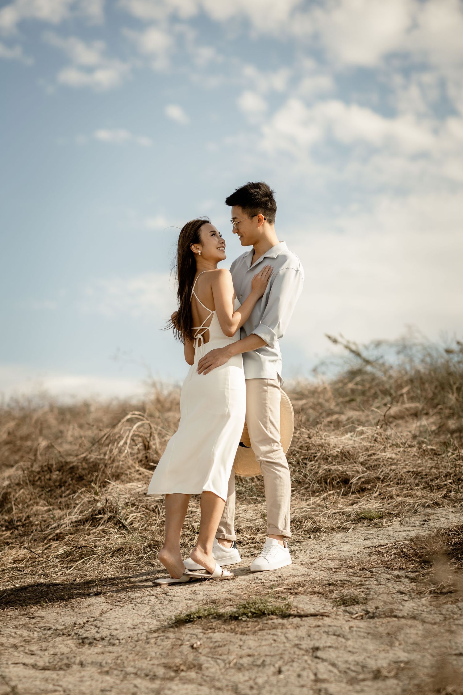 A tender moment shared between a couple in white and beige outfits on a scenic hillside path.