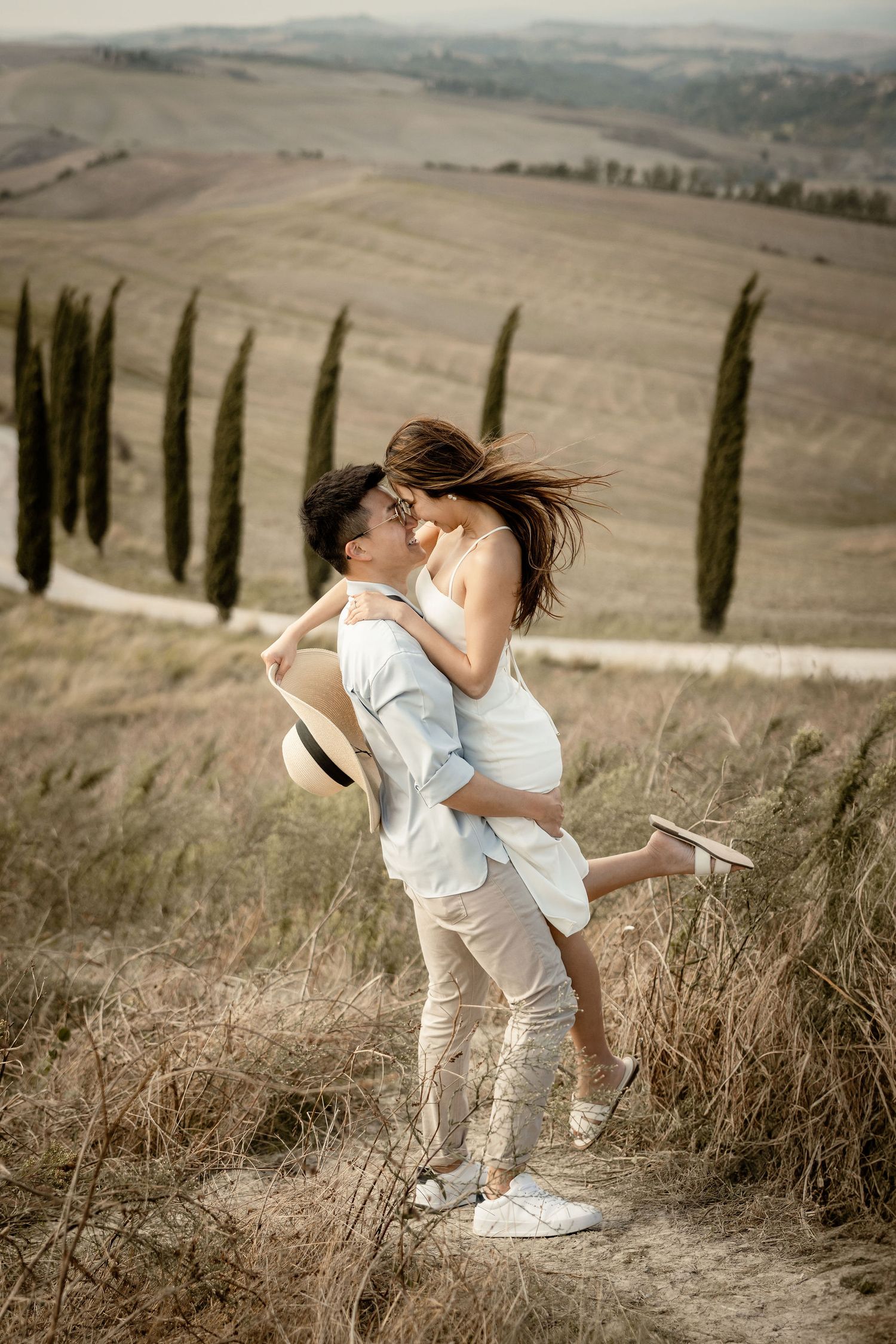 A couple in light colored clothing shares a playful moment on a scenic hillside path.