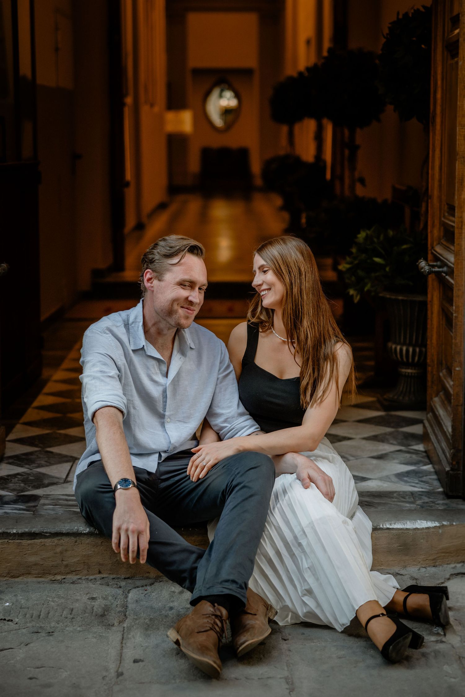 Two people share an intimate moment sitting on steps in an elegant hallway with a checkerboard floor.