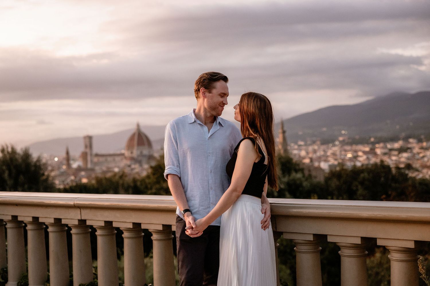 A couple shares an intimate moment overlooking Florence Italy with the Duomo cathedral visible in the distance at sunset.