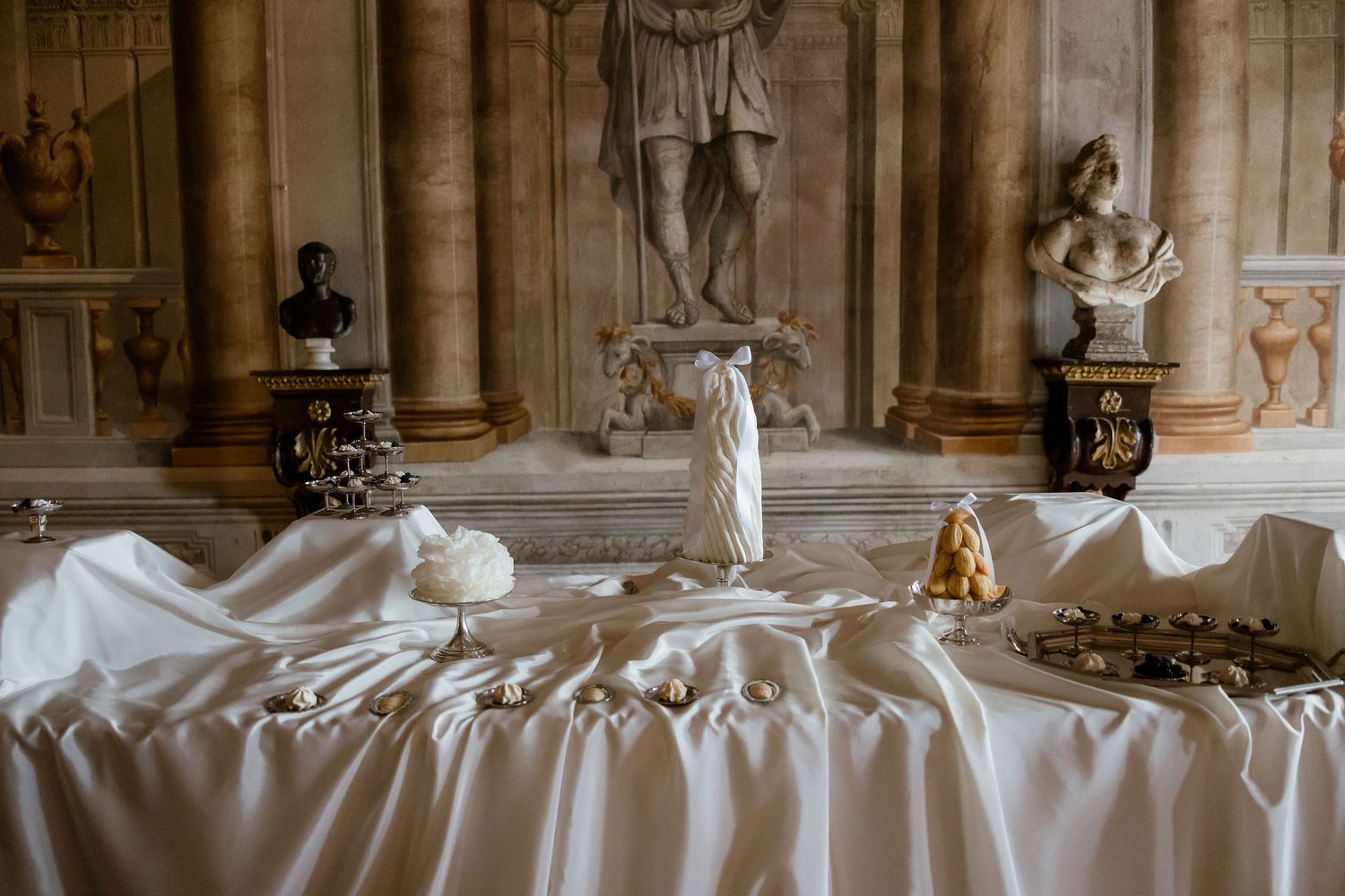 A classical interior with white draped table displaying elegant decor against stone columns and carved statues.
