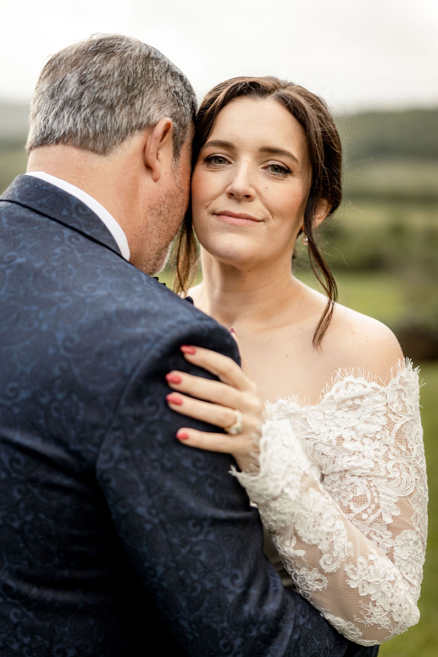 A bride and father share an emotional moment in a navy suit and white lace gown against a blurred outdoor backdrop.