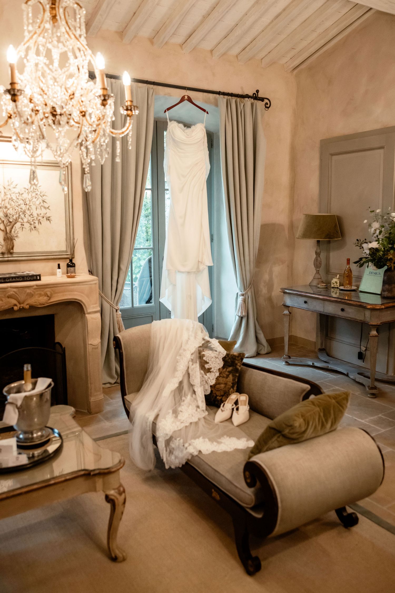 Elegant wedding dress hangs by a window in a luxurious room with crystal chandelier and antique chaise lounge.