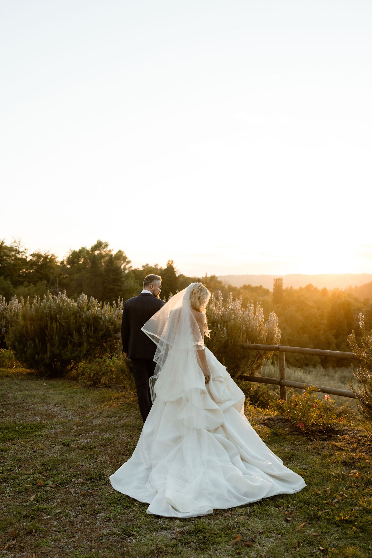 A couple in wedding attire stands together in a vineyard at sunset, with golden light illuminating their silhouettes.