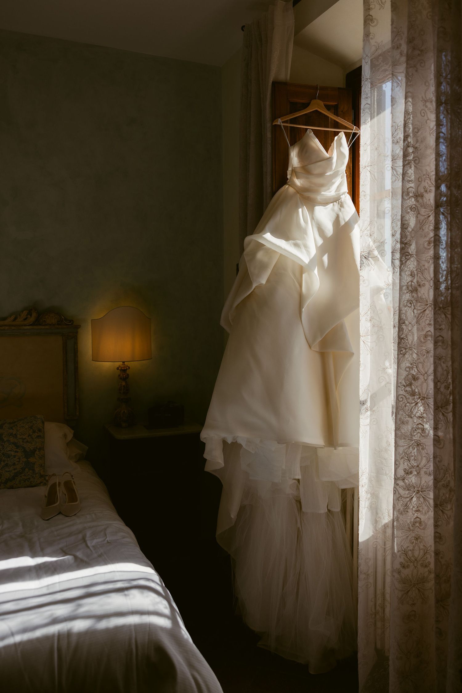 A white wedding dress hangs in a window with soft natural light casting ethereal shadows in a bedroom.