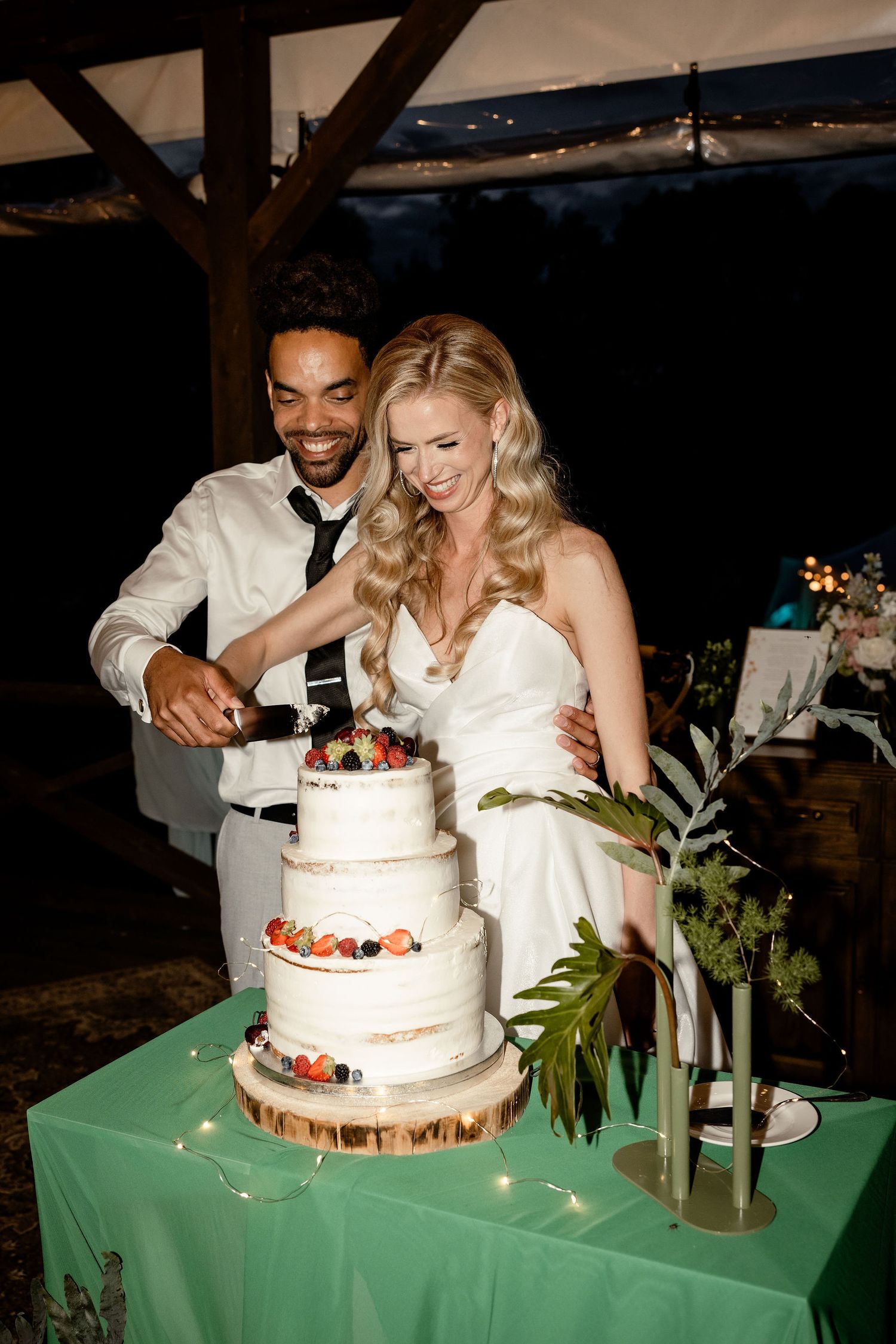 A couple cuts their white wedding cake decorated with fresh berries on a green tablecloth at their reception.