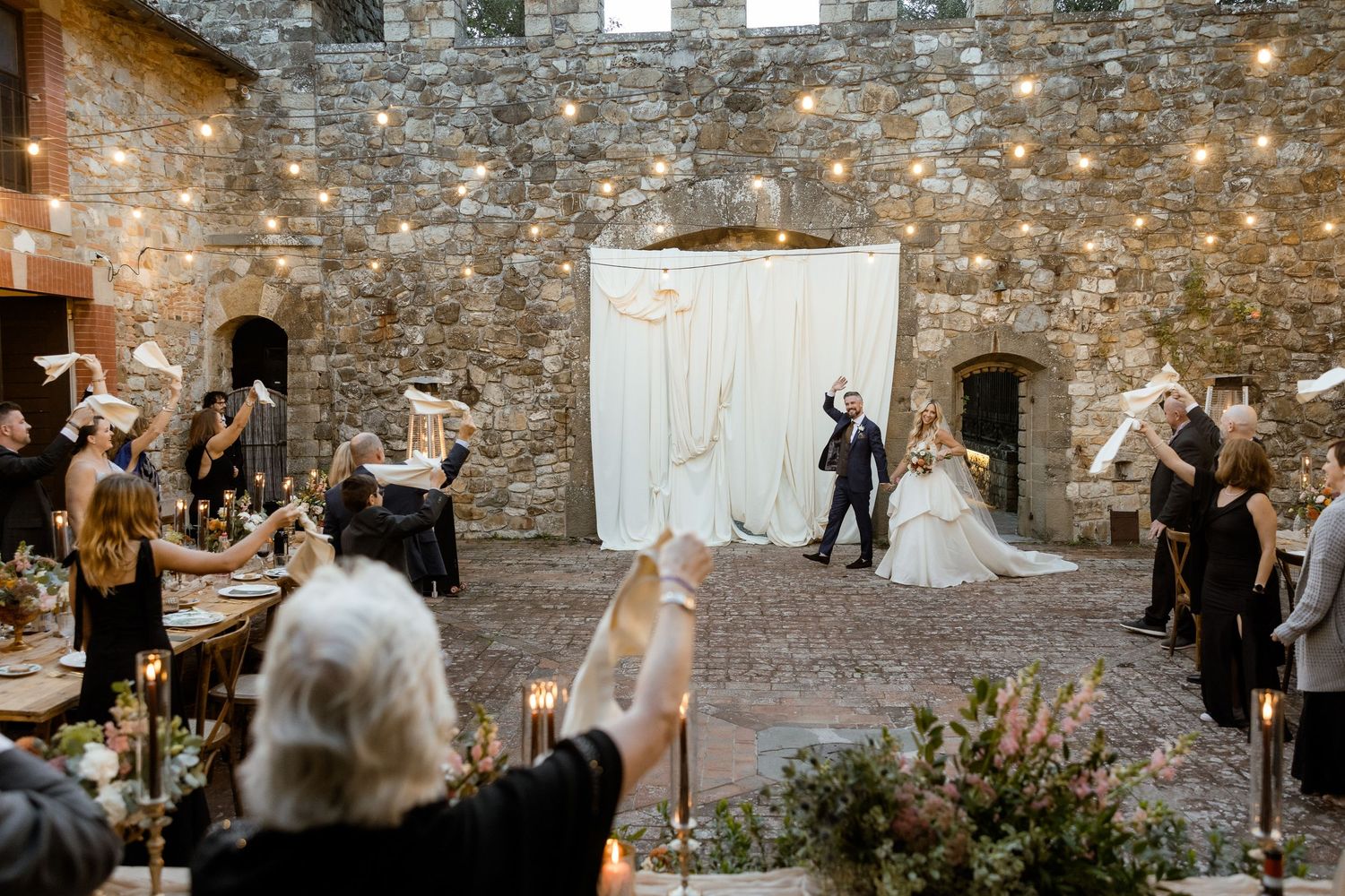 A romantic wedding ceremony takes place in a rustic stone-walled venue with twinkling string lights overhead.