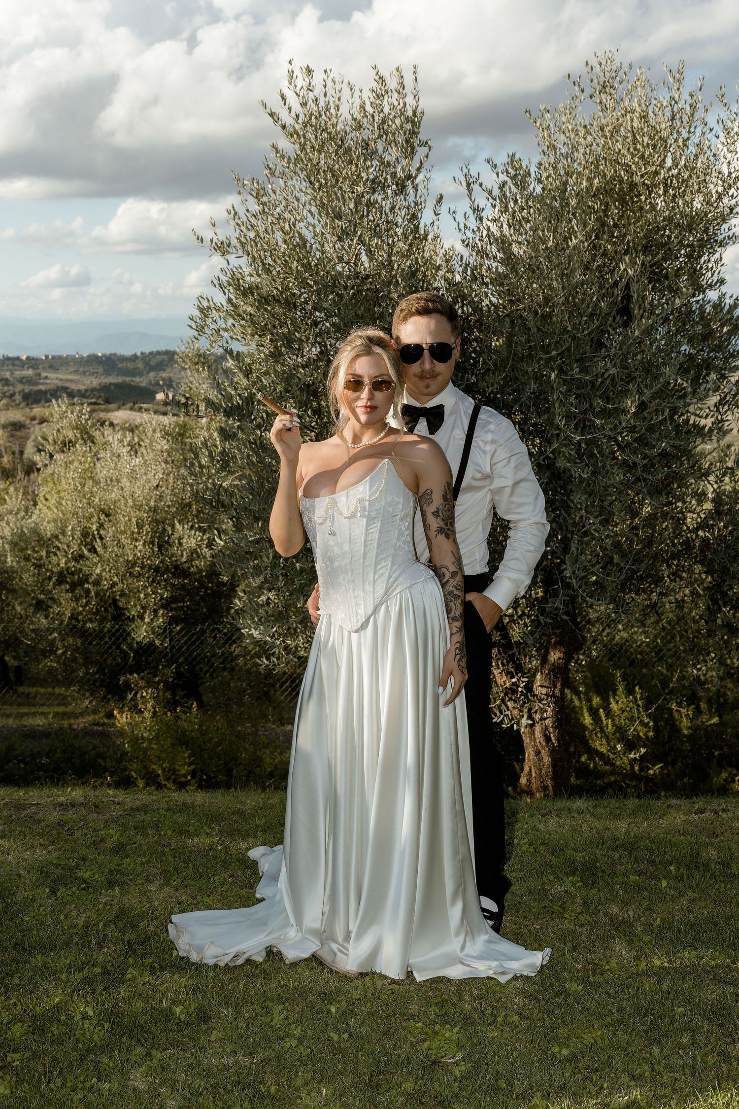 A couple in formal wedding attire poses together in an olive grove with rolling hills in the background.