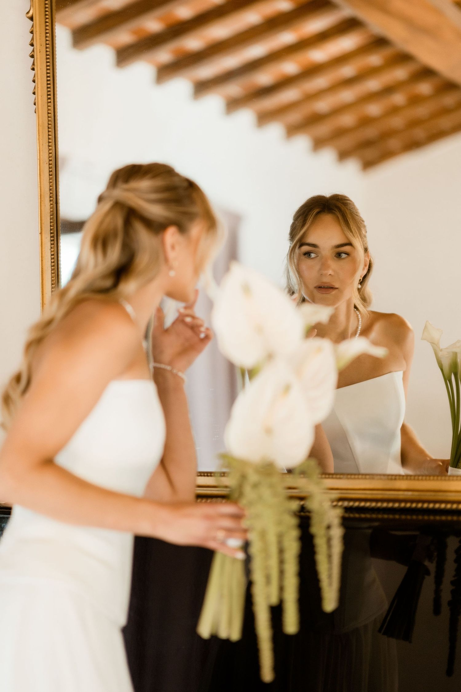 A bride in a white dress gets ready in front of a mirror with wooden beams visible overhead.
