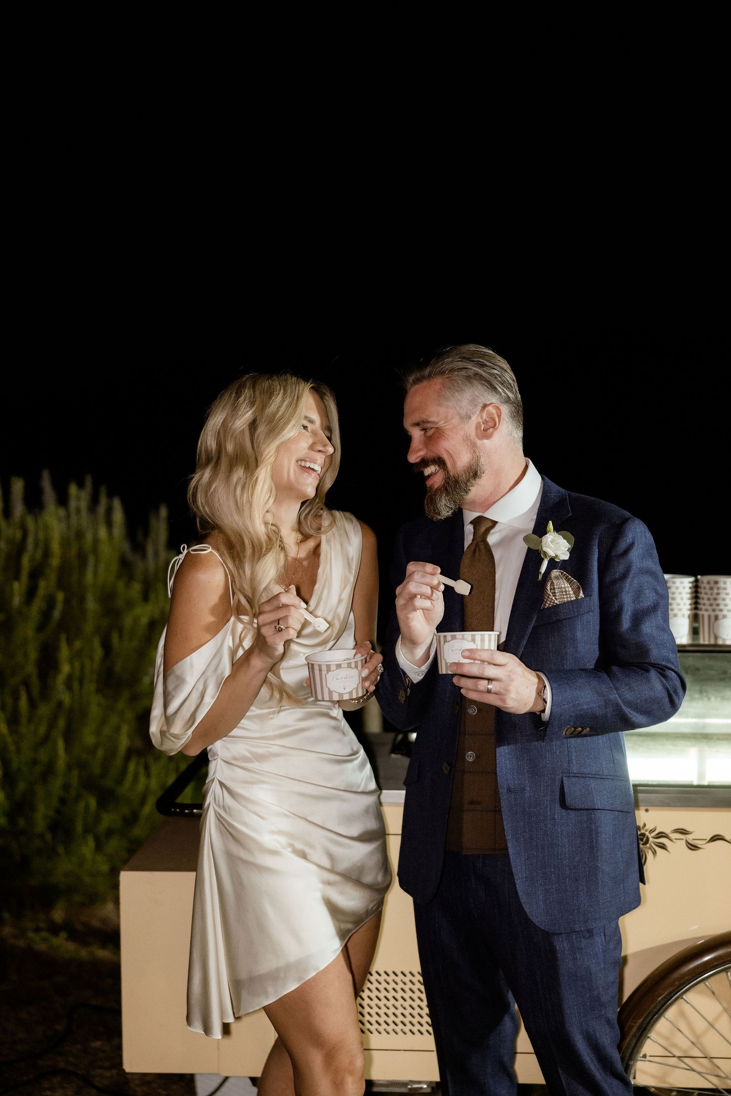 A couple in wedding attire shares a playful moment while eating cake together at their reception.