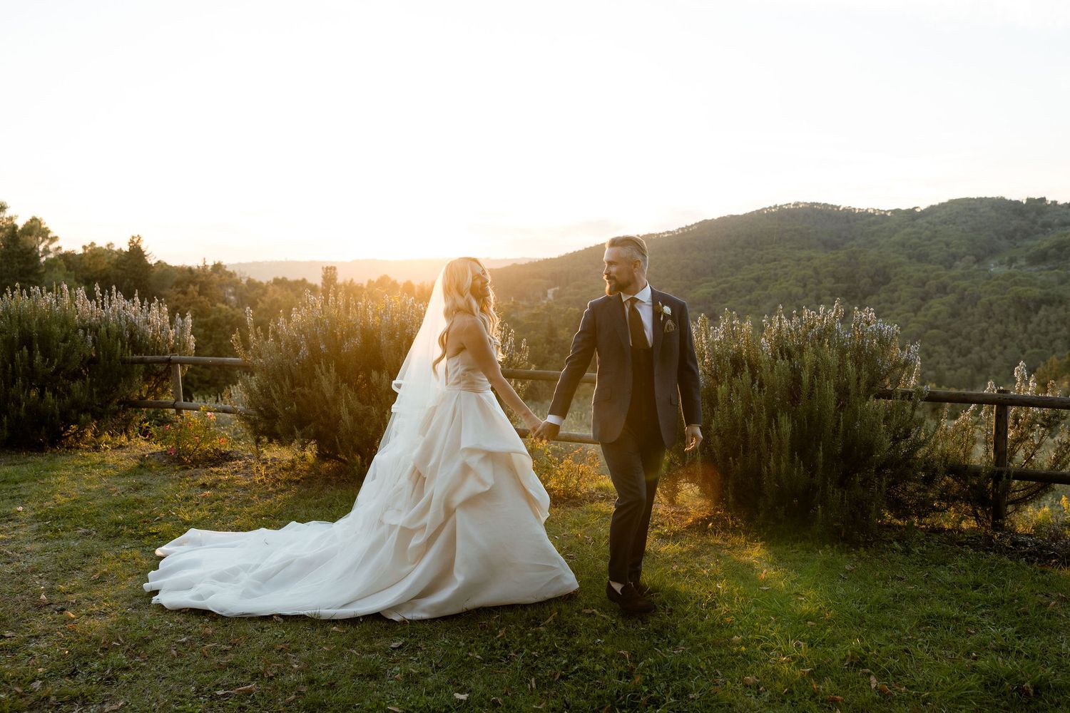 A couple in wedding attire walks together on a hilltop at golden hour with mountains in the background.