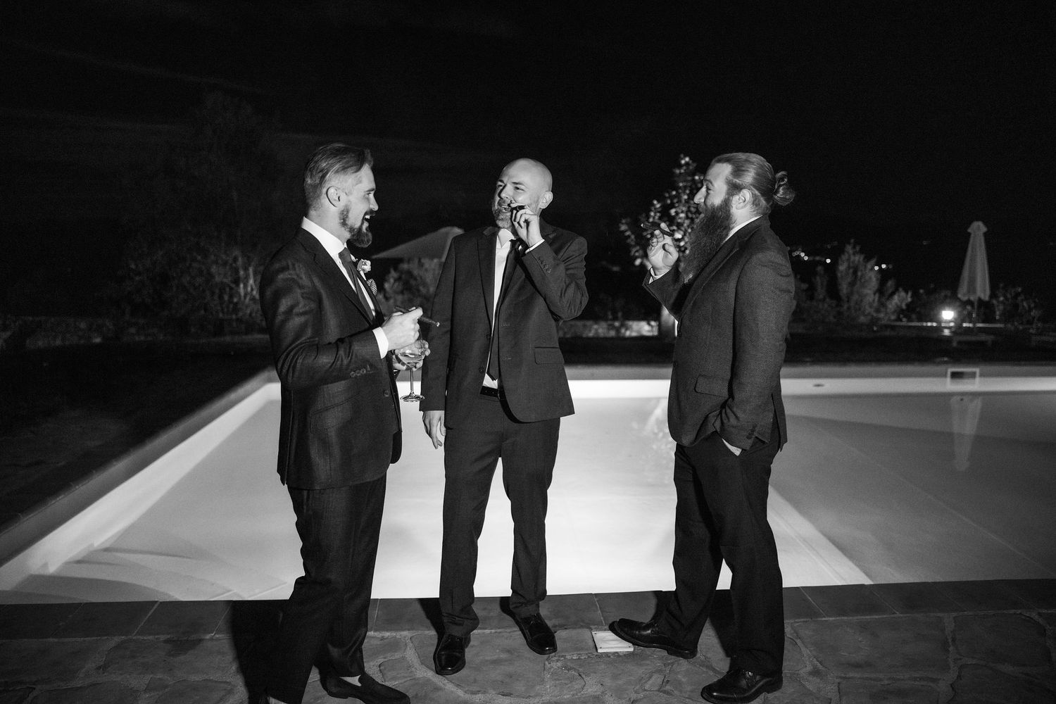 Three people in formal black suits having a conversation while standing outdoors at night by a pool.