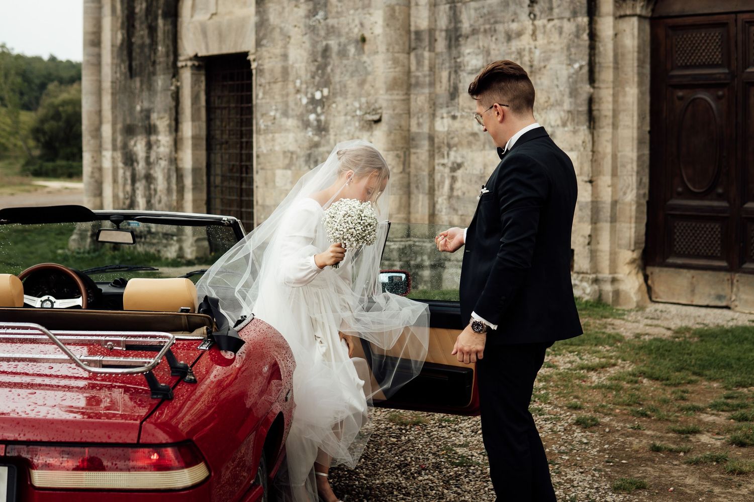 A couple shares a tender moment beside a vintage red car outside a historic stone church on their wedding day.