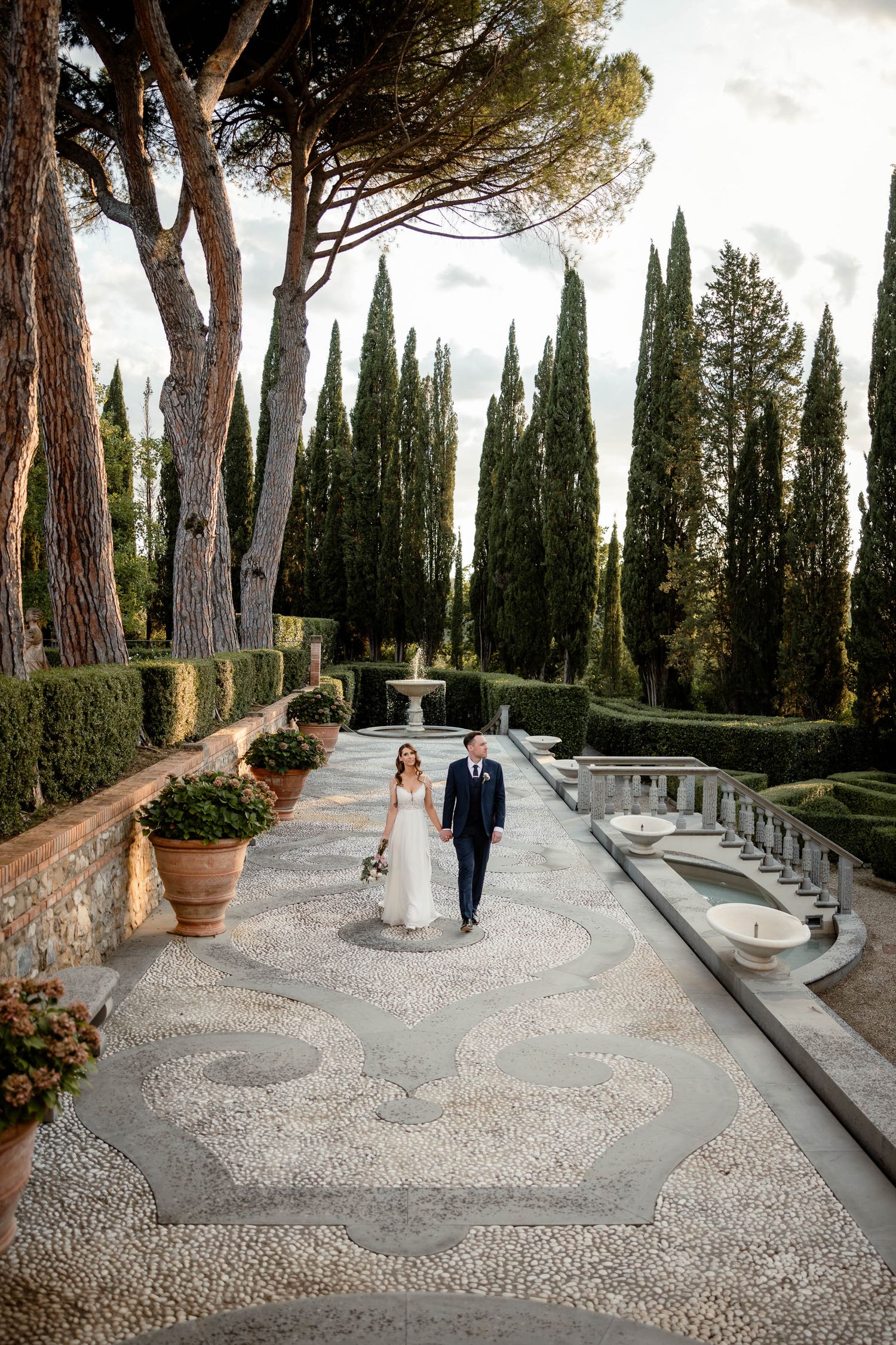 Elegant couple walking stone mosaic path lined with tall cypress trees in formal Italian garden.