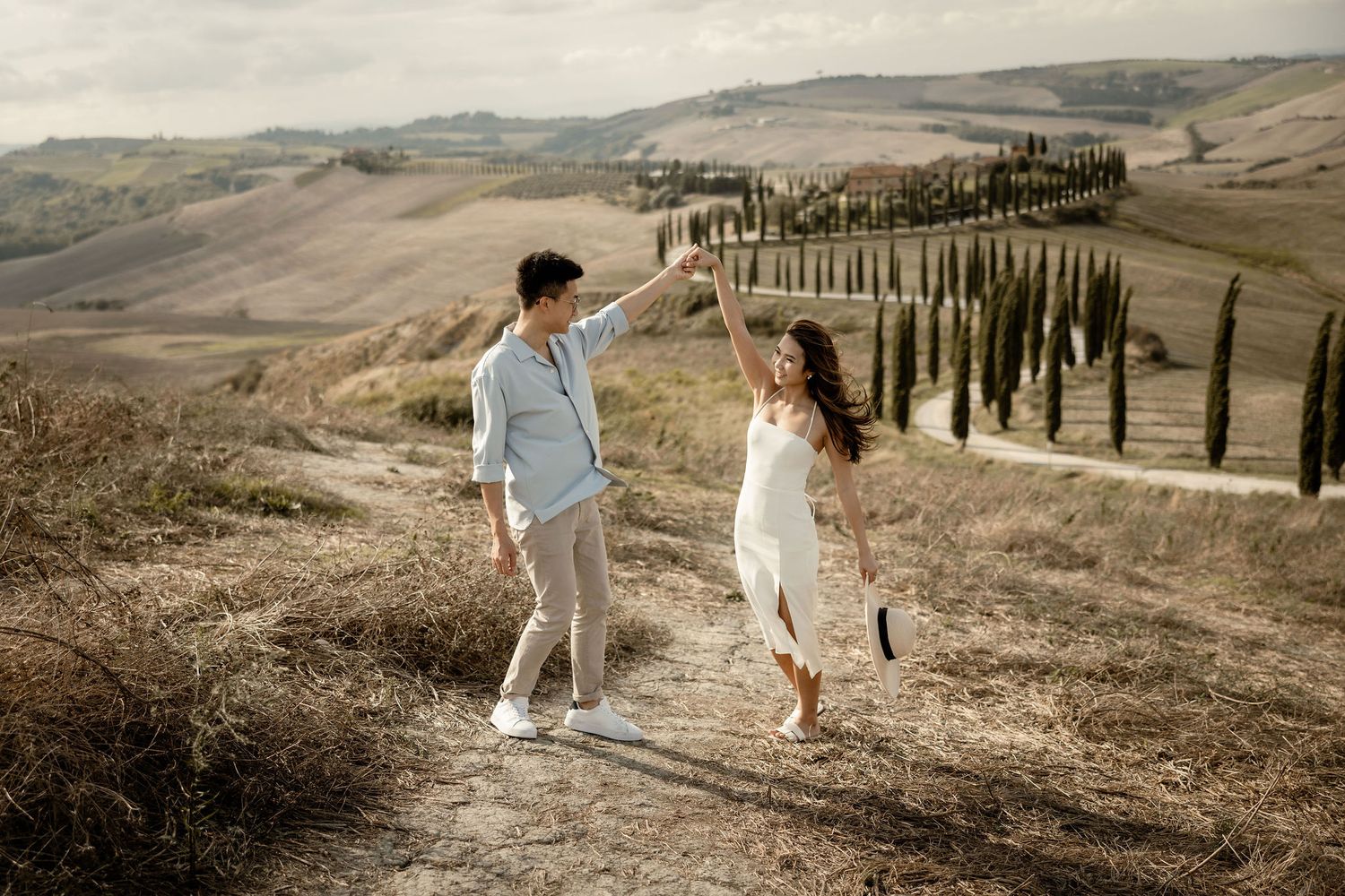 A couple walks hand in hand on a Tuscan hillside lined with cypress trees in elegant casual wear.