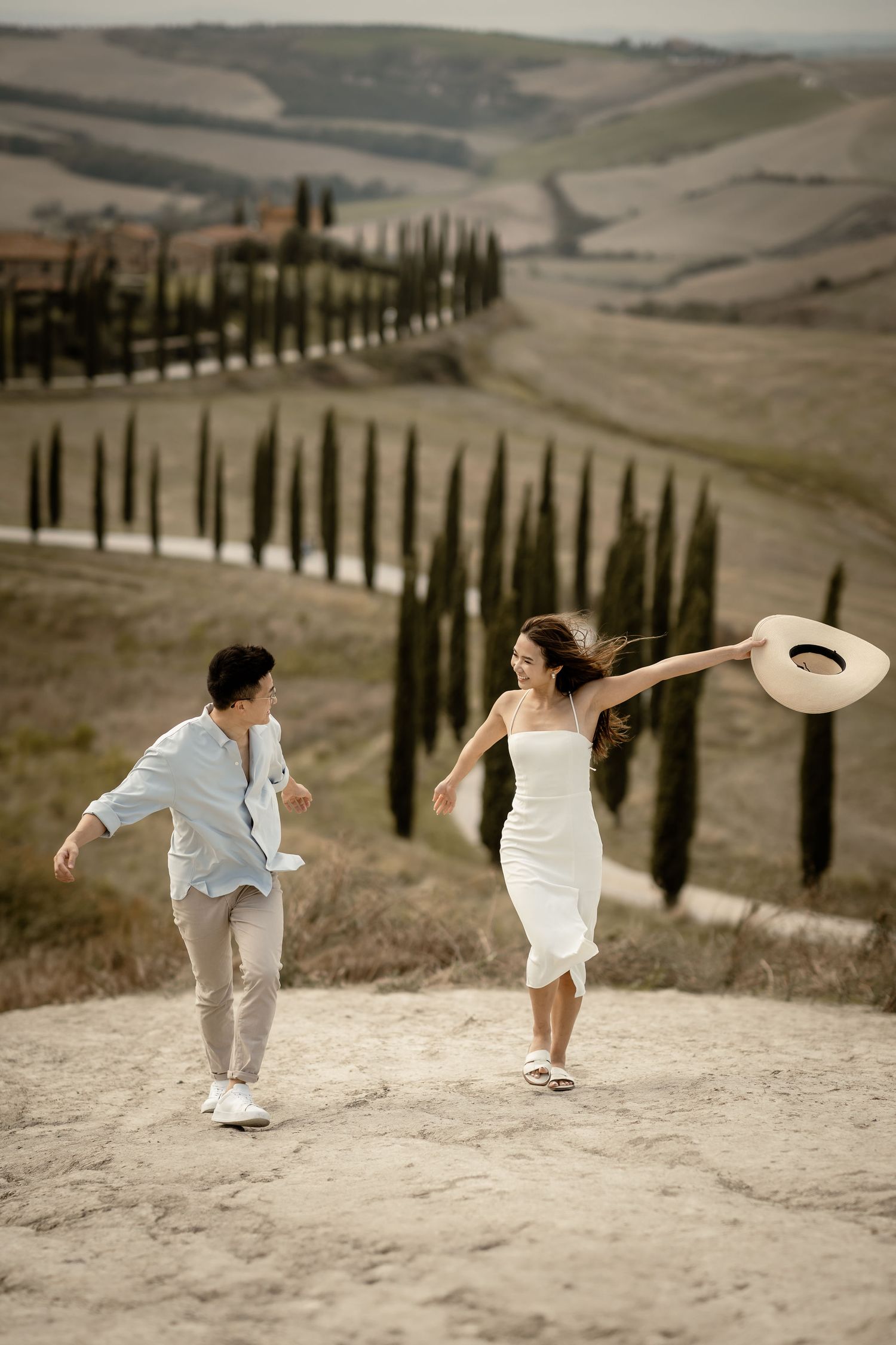 Joyful couple dancing on dirt path with iconic Tuscan cypress tree landscape.