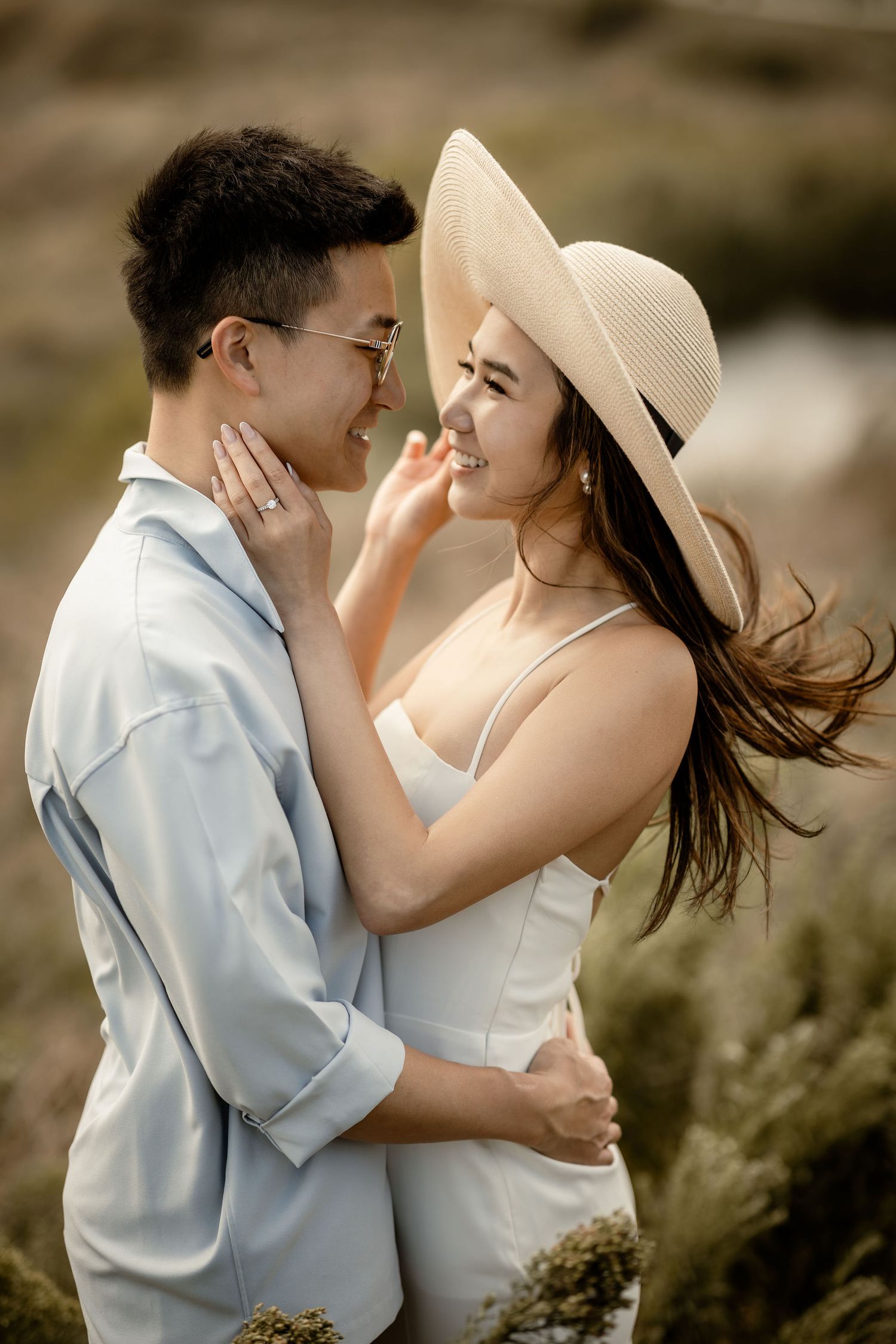 Tender moment between couple in white attire with sun hat against natural backdrop.