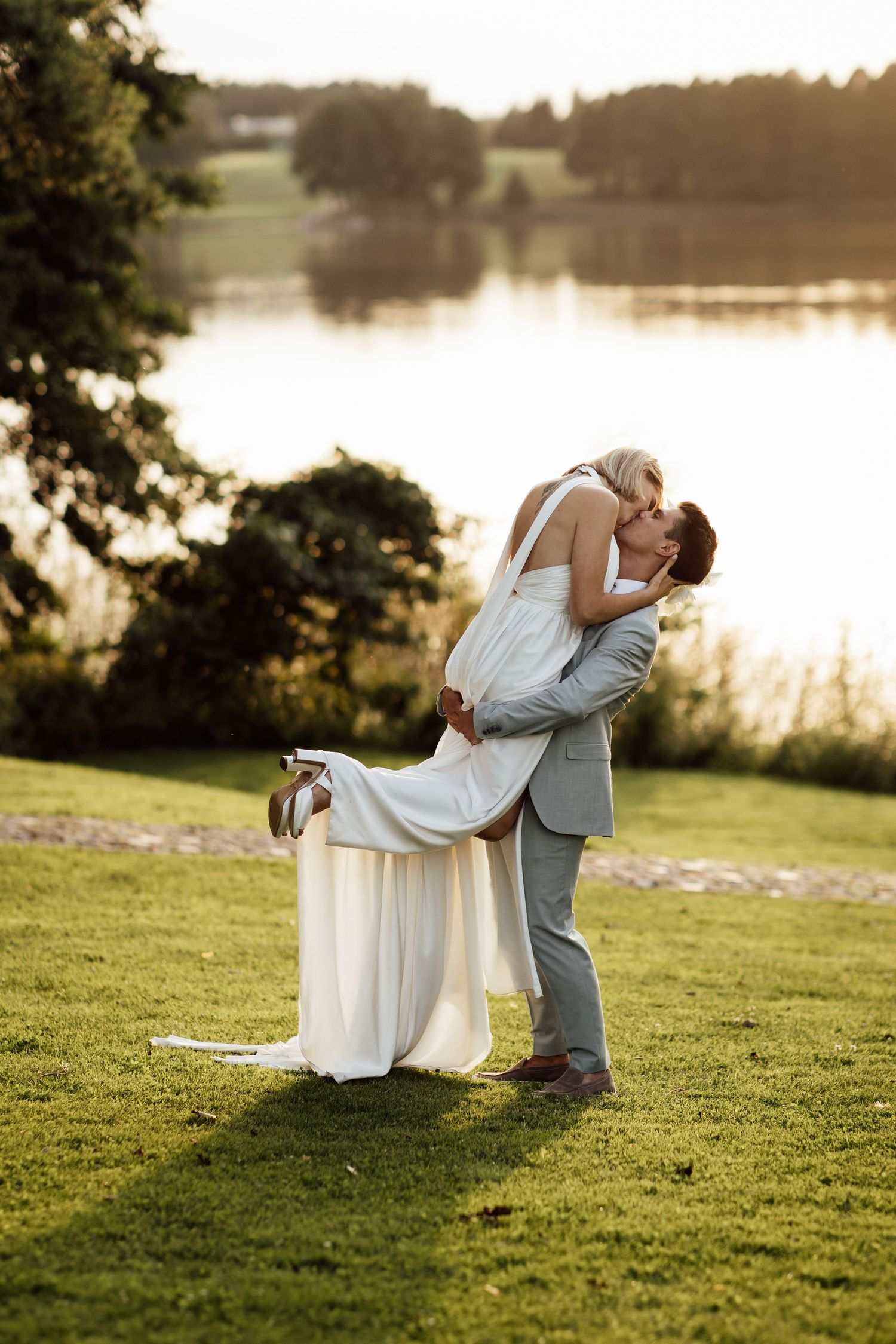 Romantic couple embraces during sunset by a lake, with lush green grass and golden light creating a dreamy atmosphere.