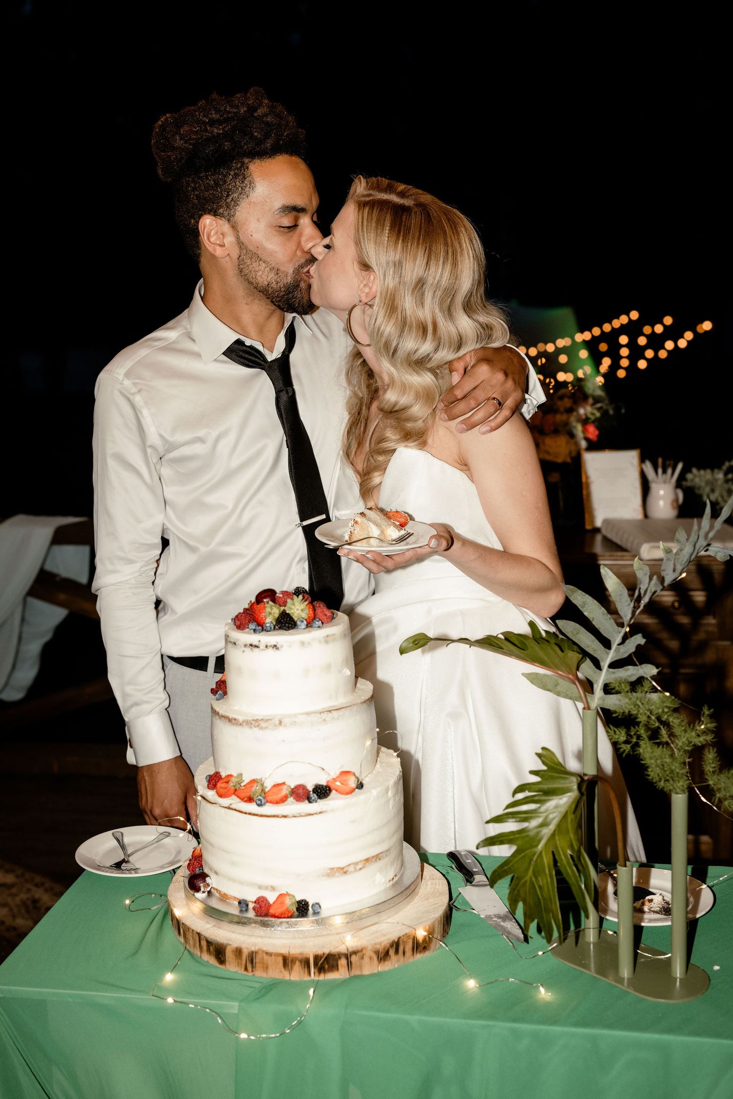 A couple shares a kiss next to their three-tiered white wedding cake decorated with berries on a green tablecloth.