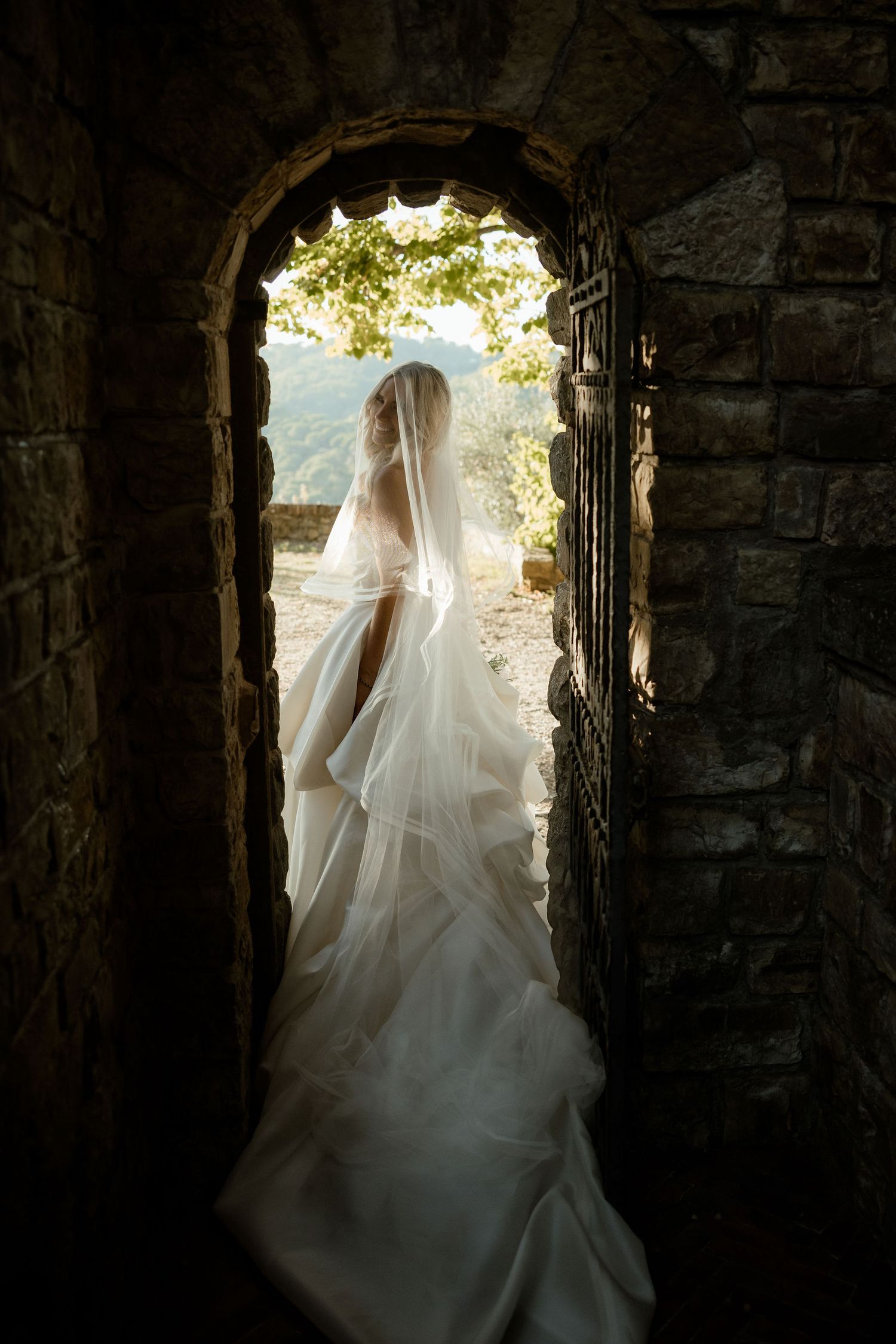 A flowing white wedding dress silhouetted in a stone archway doorway with sunlight streaming through the opening.