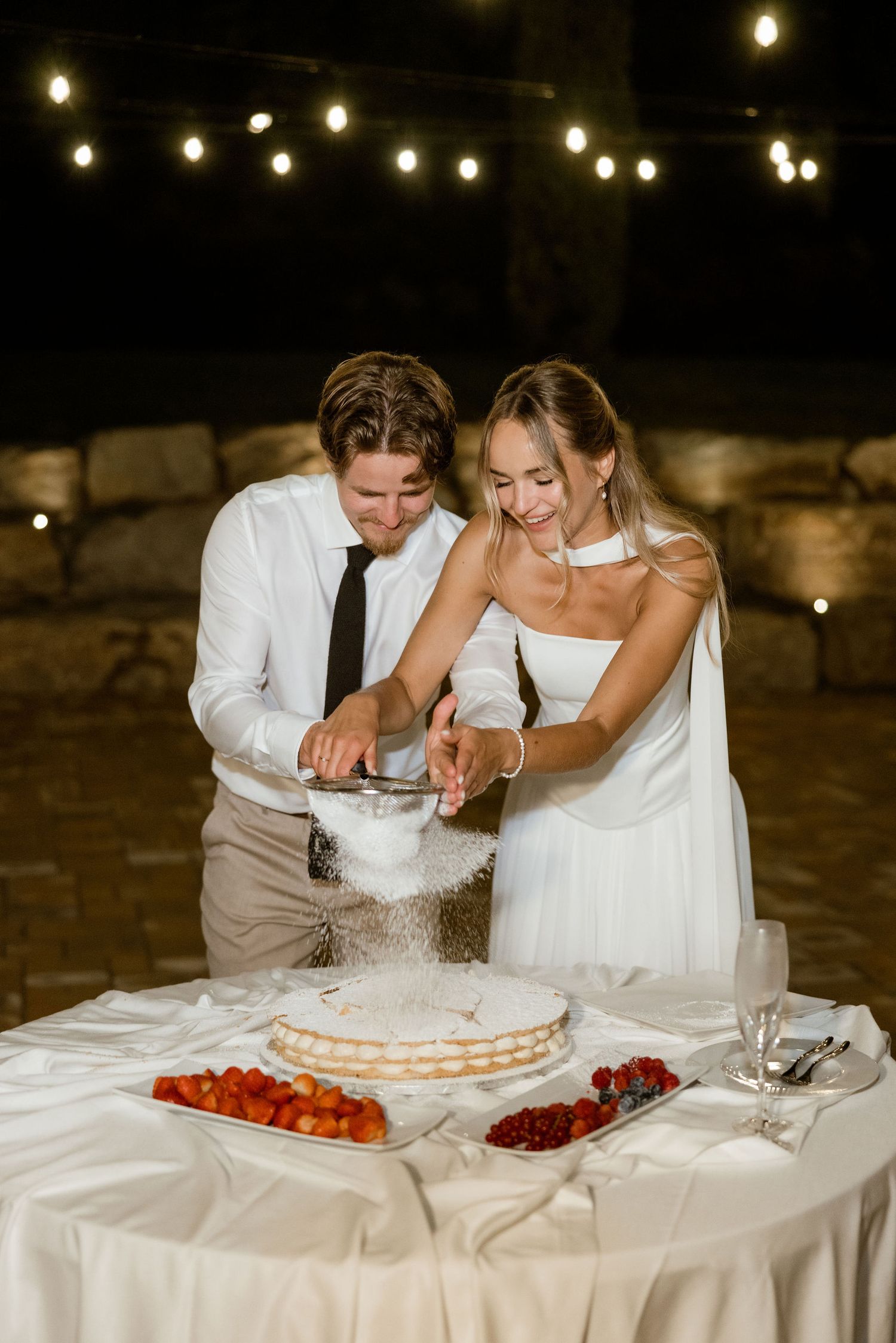 A couple cuts into a traditional white wedding cake together while surrounded by string lights and fruit decorations.