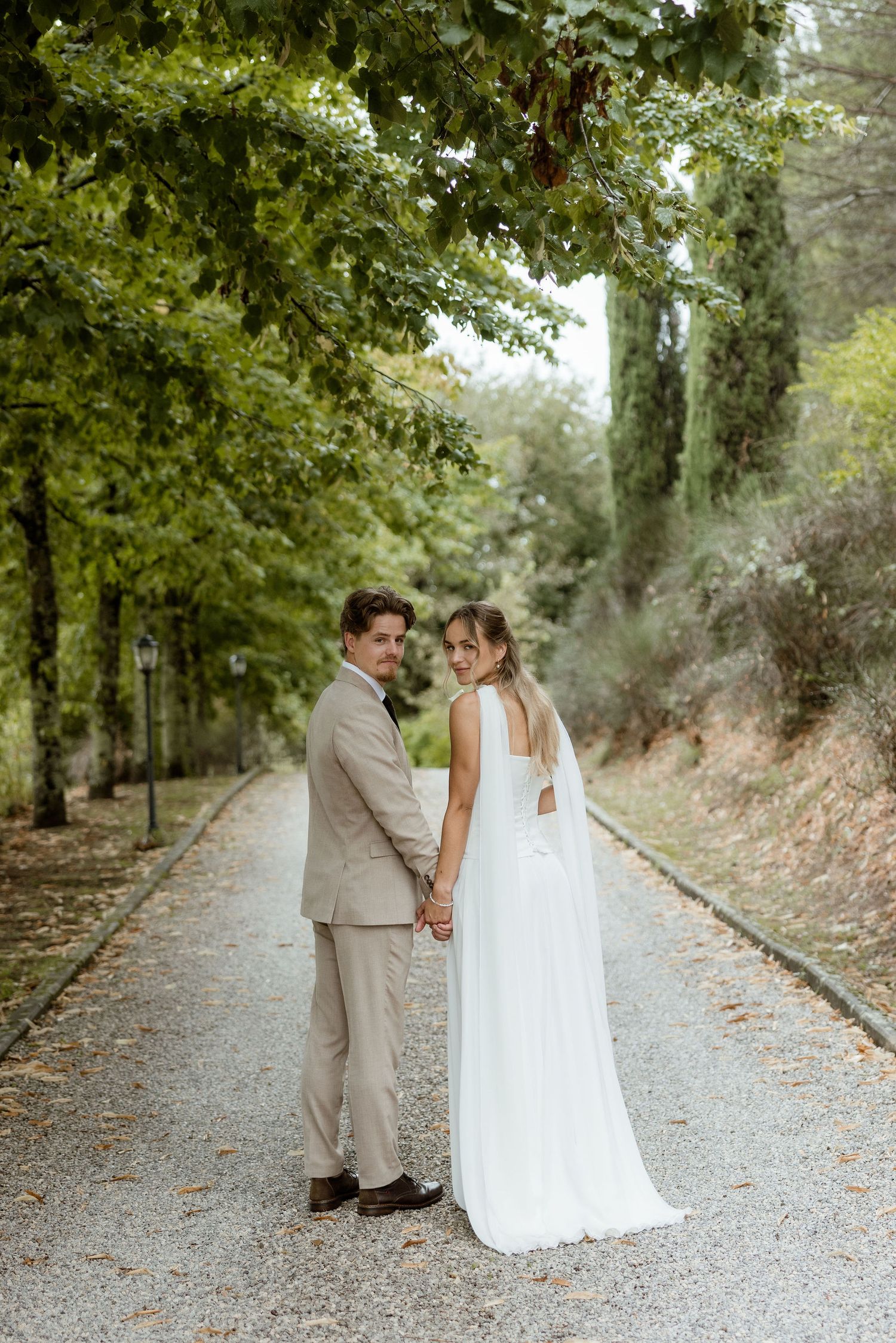 A couple in wedding attire shares intimate moments on a tree-lined gravel path during their outdoor photo session.