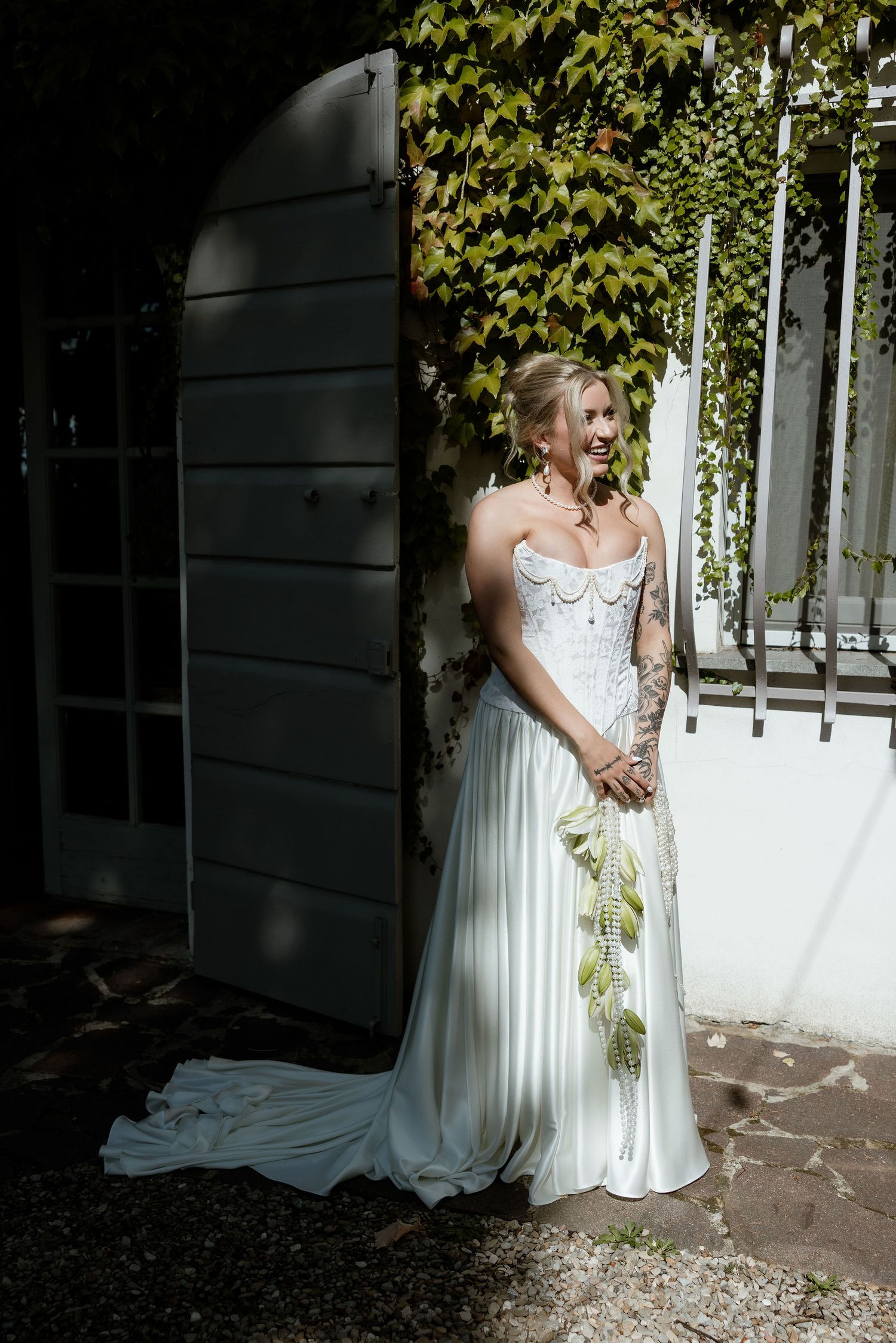 Two people in white wedding attire embrace under green foliage on a sunny outdoor patio.
