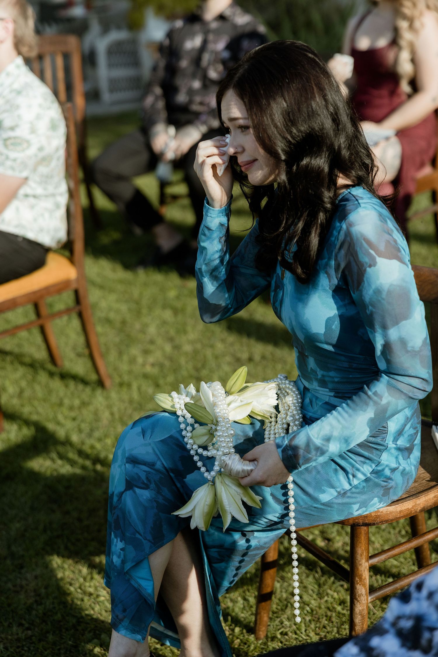Person in a flowing blue dress sits on a wooden chair outdoors holding white flowers in sunlit garden setting.