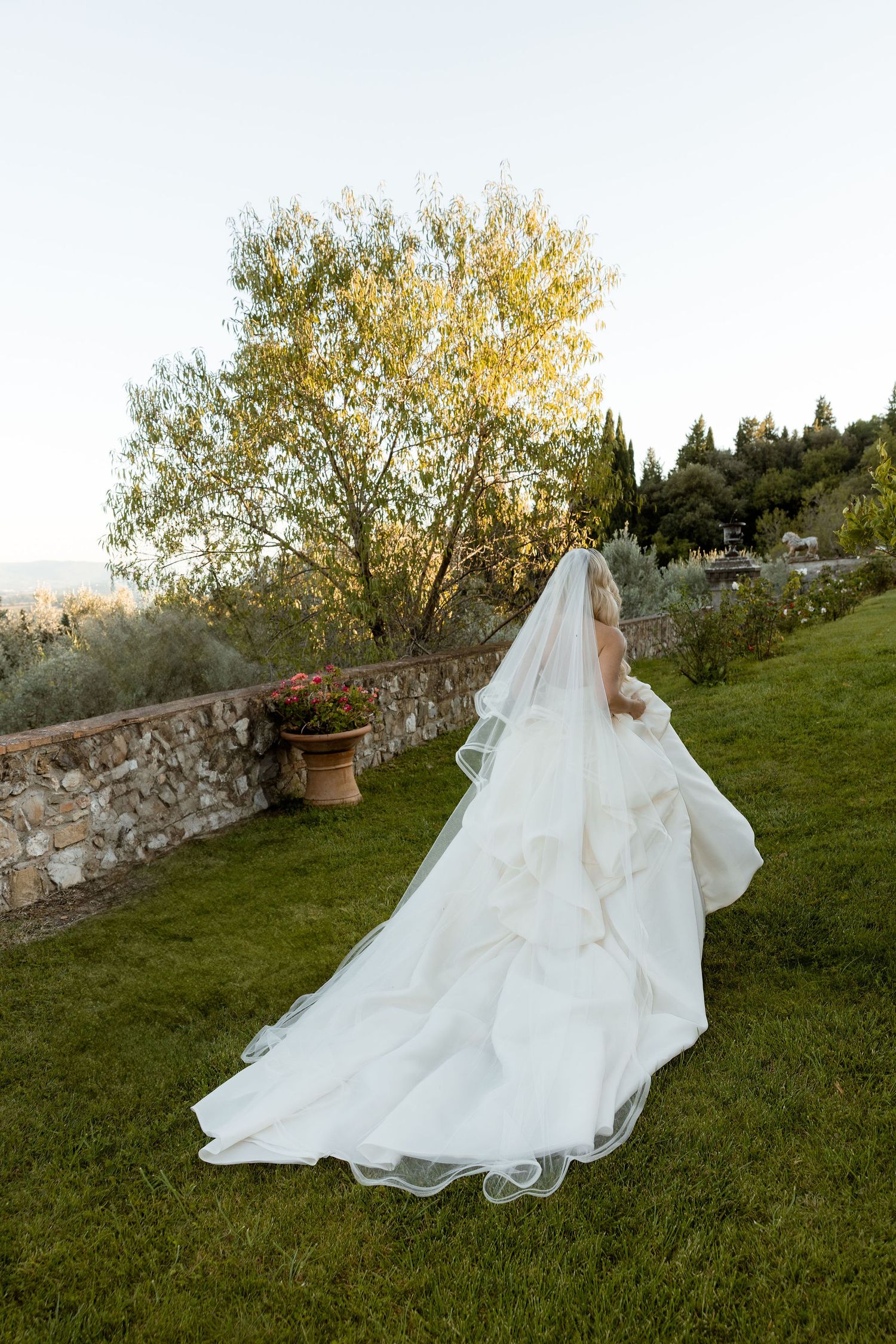 A flowing white wedding dress with a long train and veil trails across a grassy hillside garden setting at sunset.