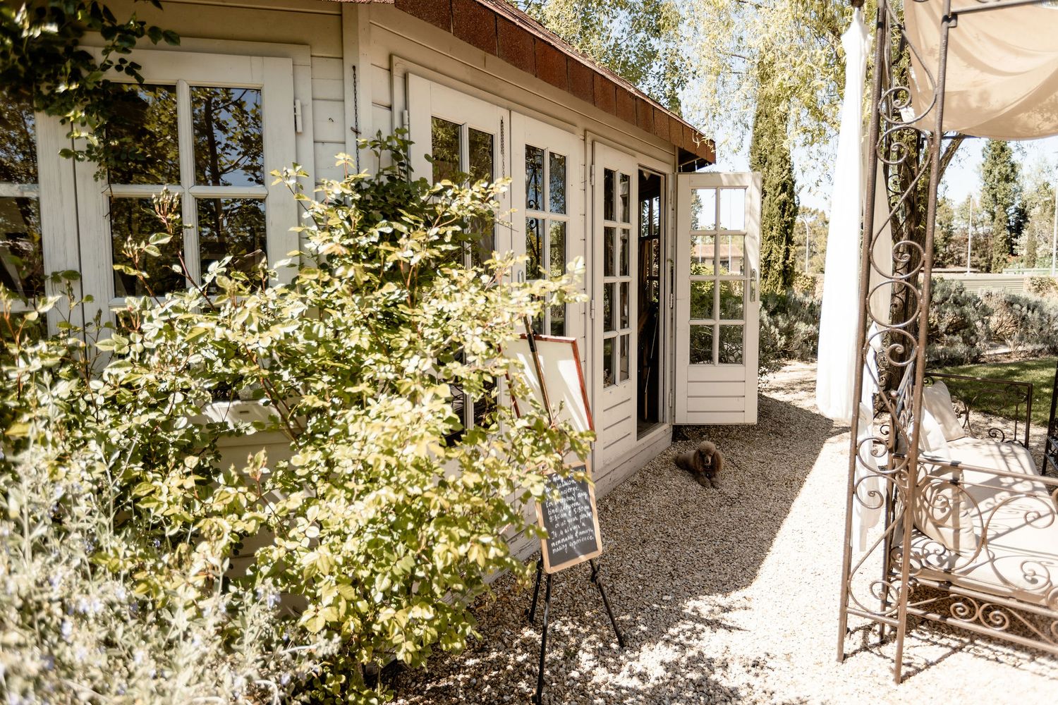 Charming white cottage exterior with French doors and lush greenery along a gravel path in warm sunlight.