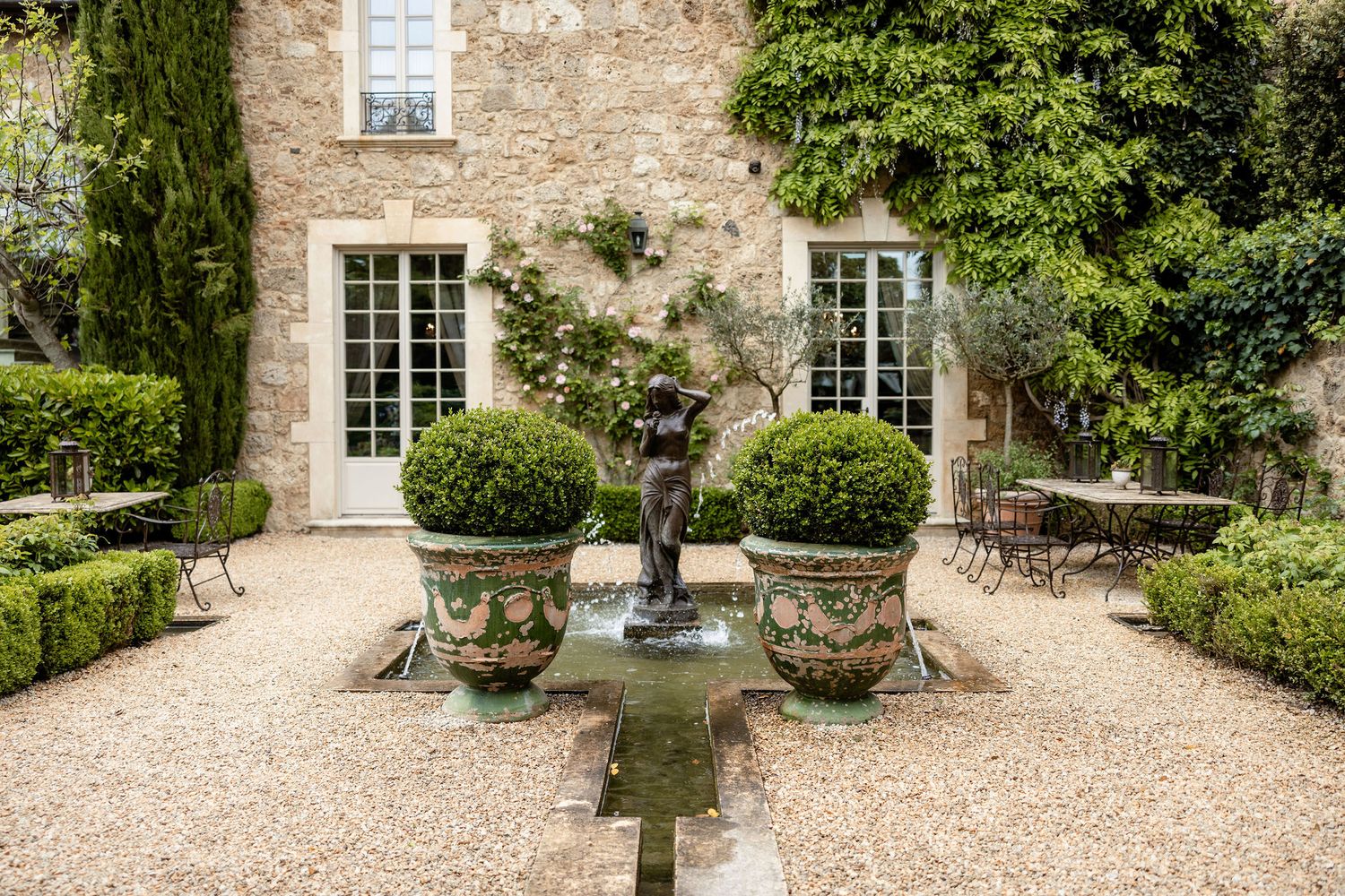 A stone path garden courtyard with multiple circular planters and white framed windows on a rustic stone building.