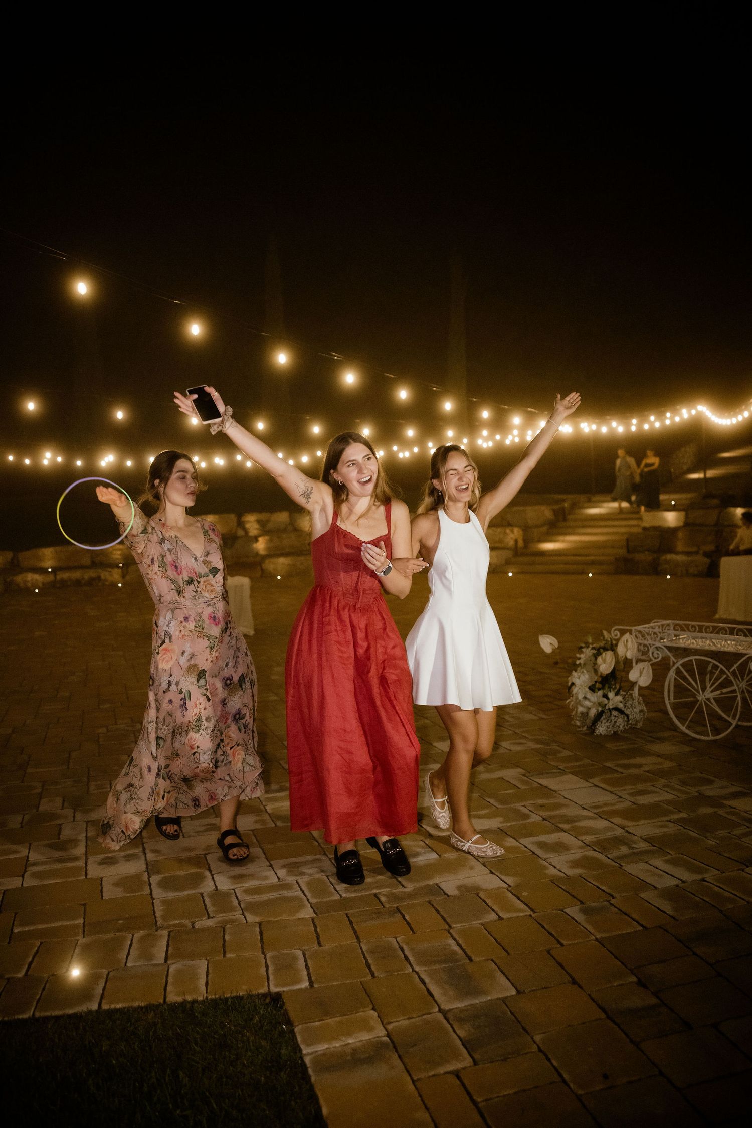 Three women dancing joyfully at night under string lights on a brick patio during an outdoor celebration.