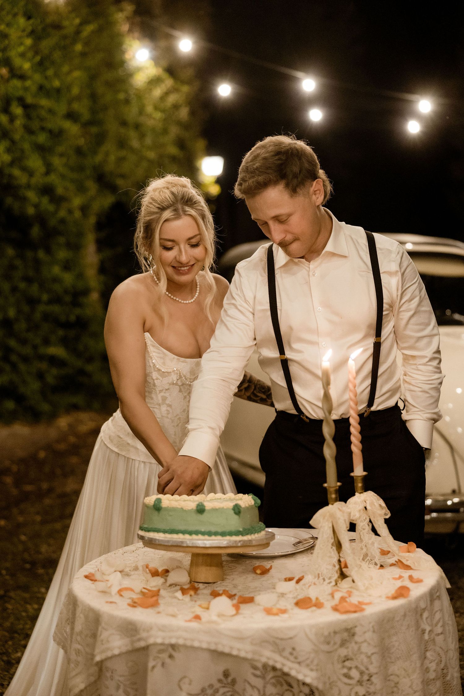 A couple cuts their wedding cake together under string lights at an outdoor evening reception.