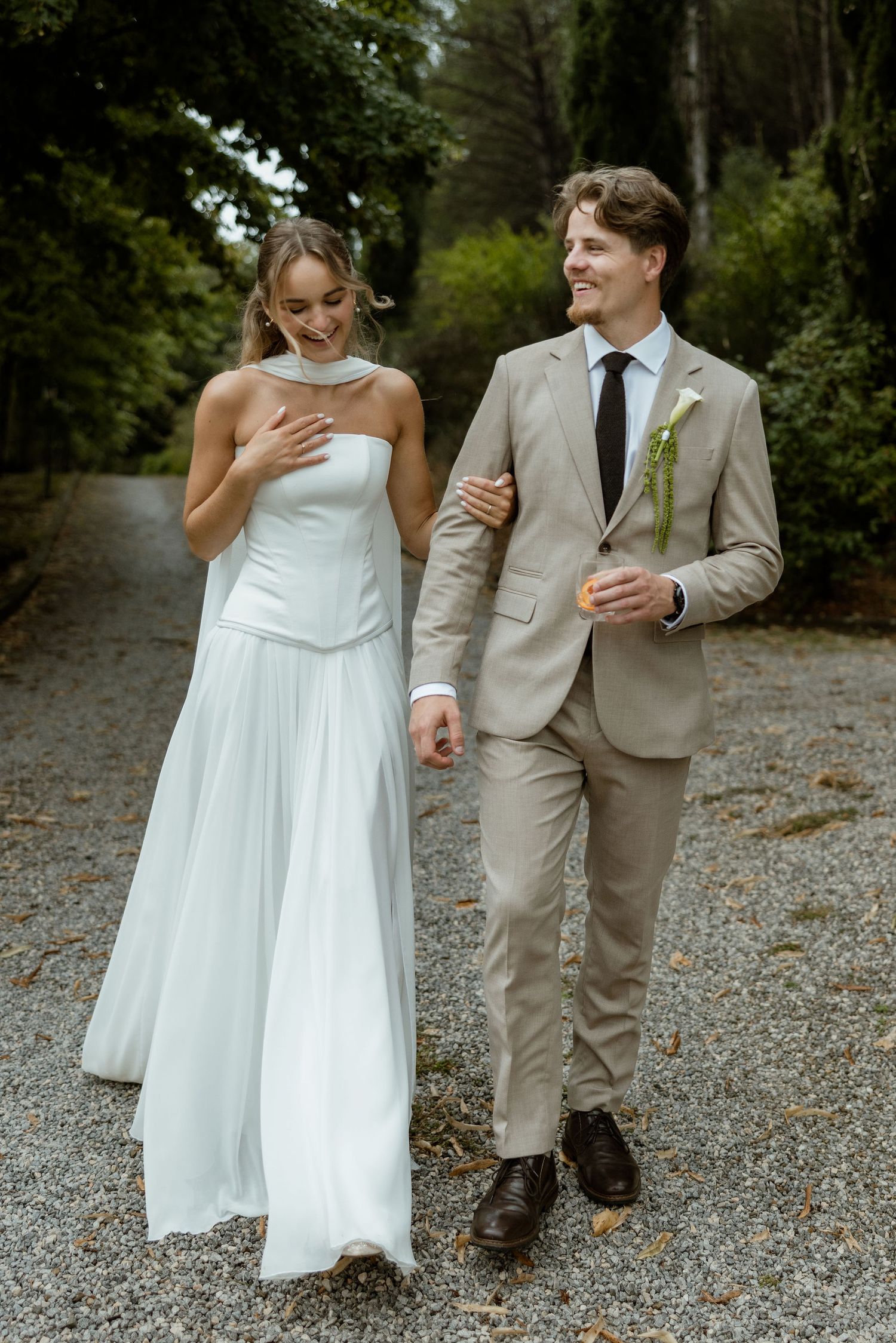 A couple in wedding attire walking and laughing together on a gravel path surrounded by lush greenery.