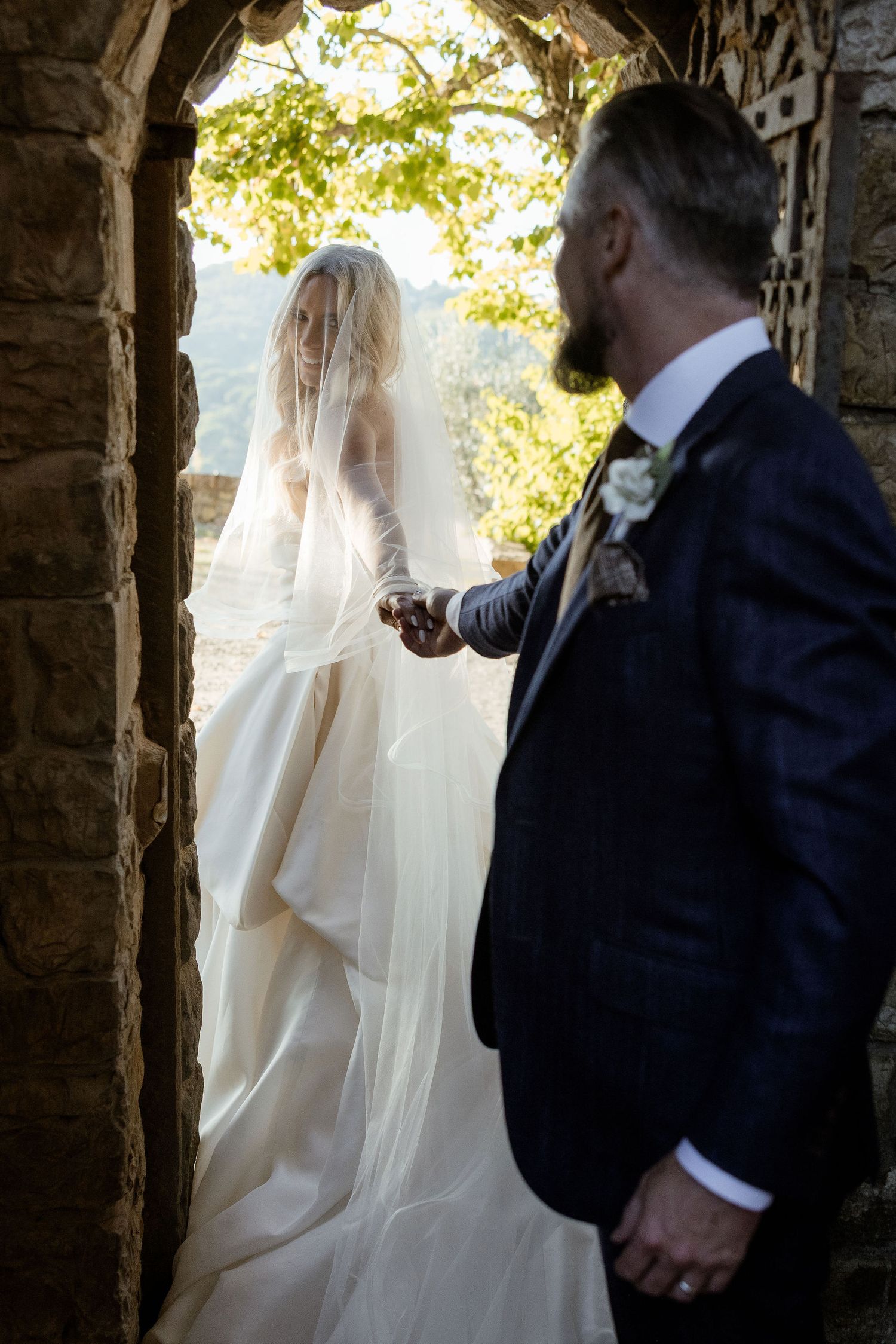 A bride and groom share an intimate moment in a stone archway with natural sunlight filtering through.