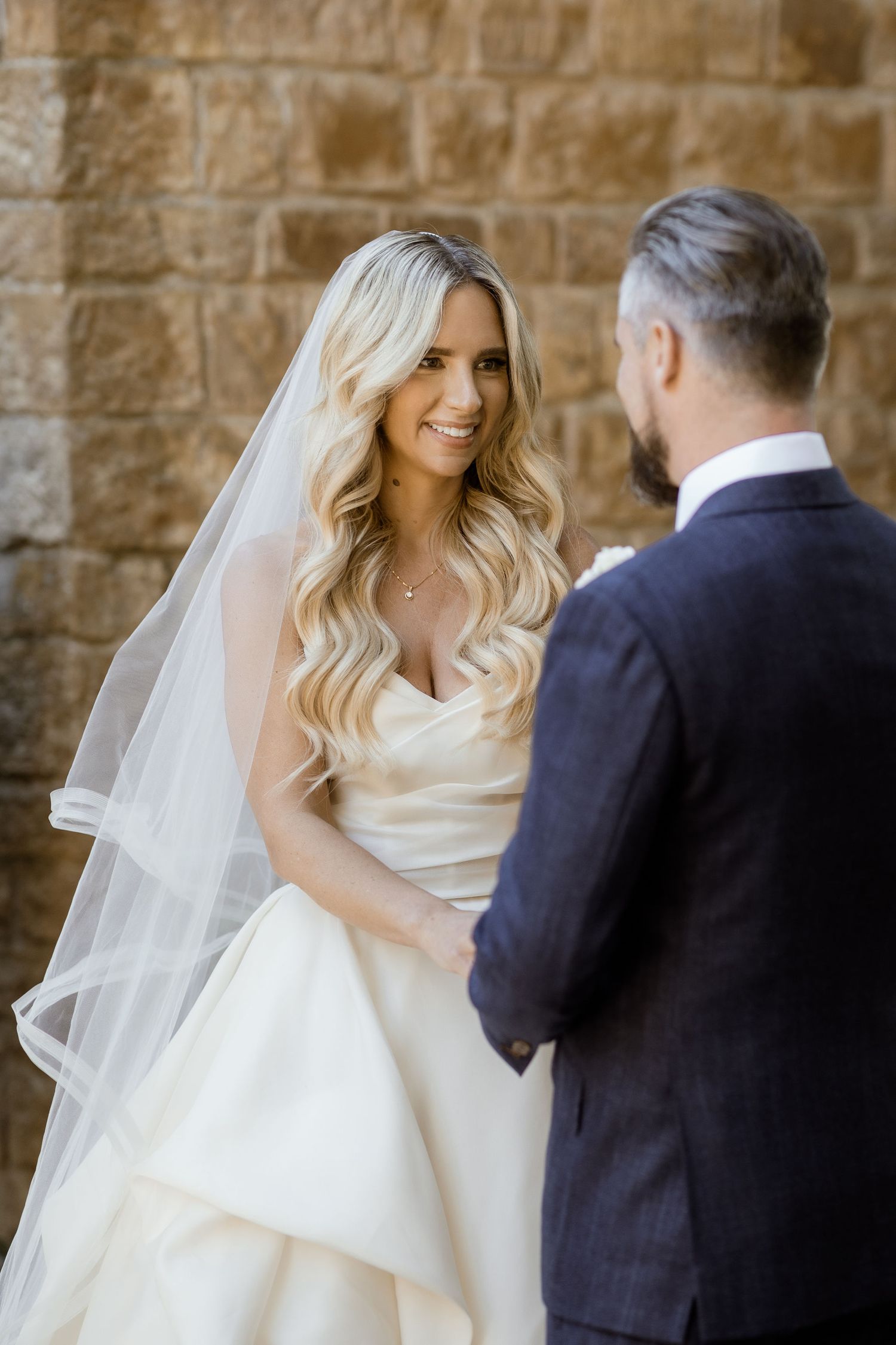 A bride in a white layered wedding gown with flowing veil stands with her groom against a stone wall.