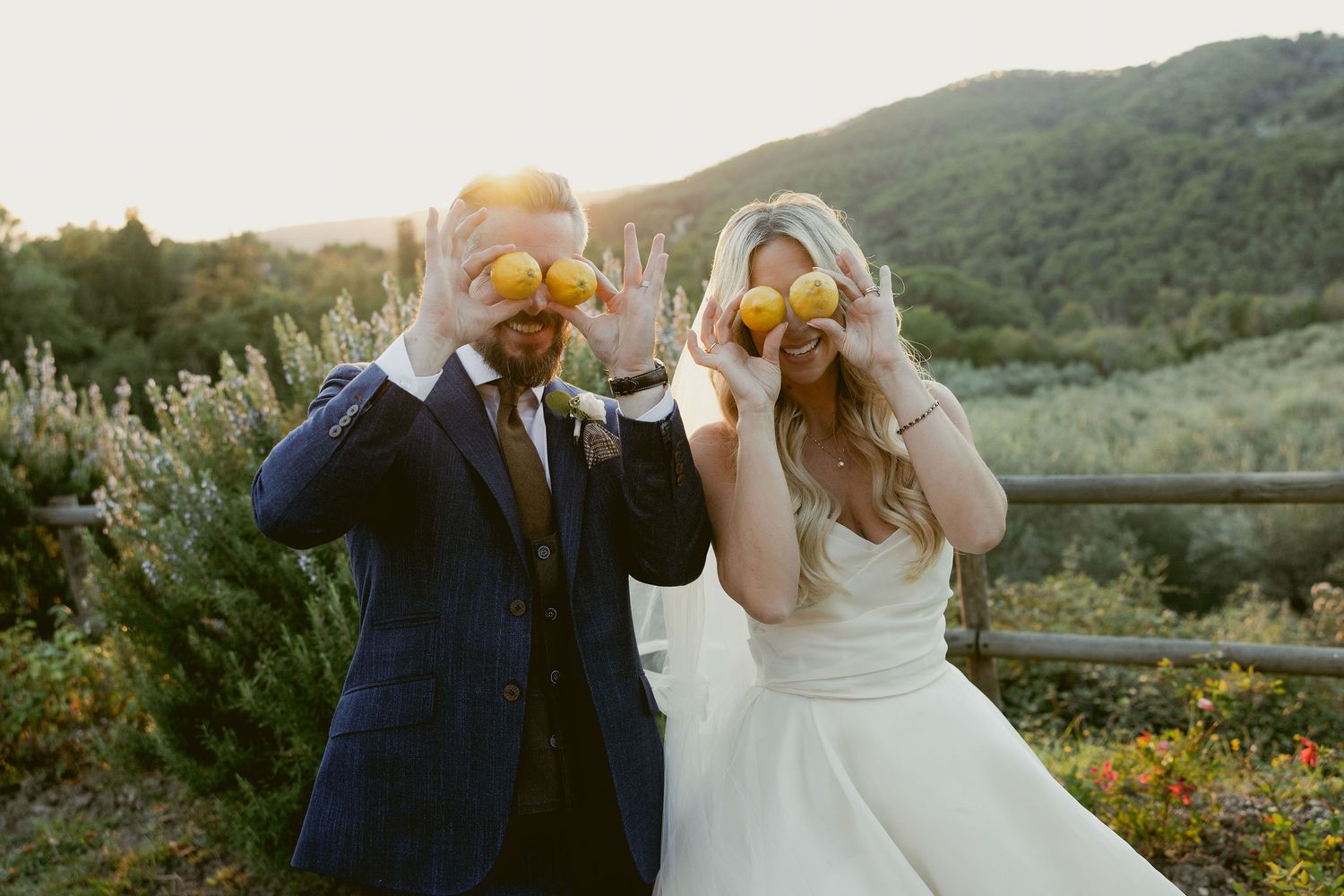 A romantic outdoor wedding scene at sunset with a couple embracing on a hillside overlooking a scenic landscape.