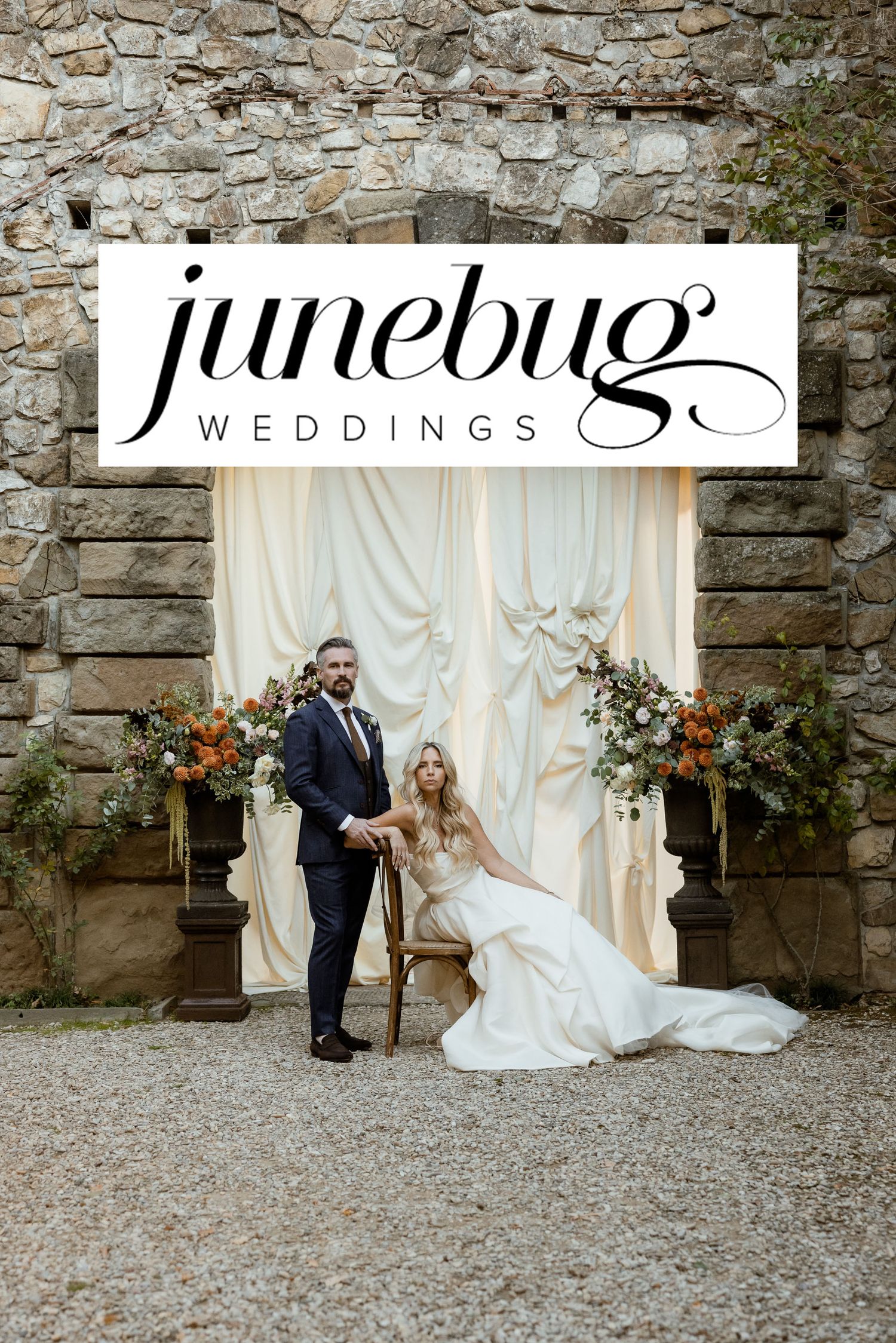 Wedding couple poses elegantly against rustic stone wall backdrop with white drapes and autumn floral arrangements.