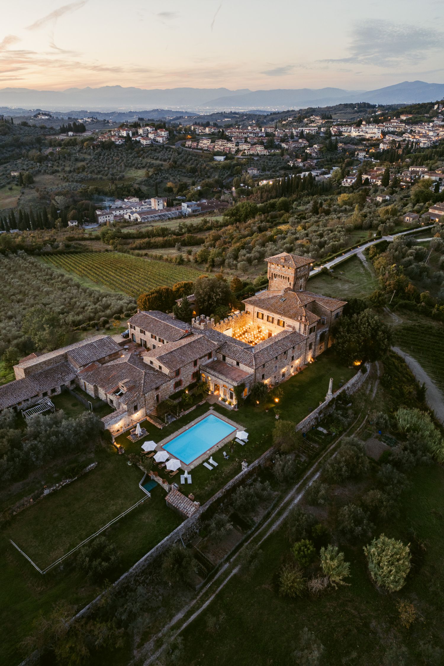 Aerial view of a luxurious Italian villa with swimming pool surrounded by lush gardens and countryside at sunset.