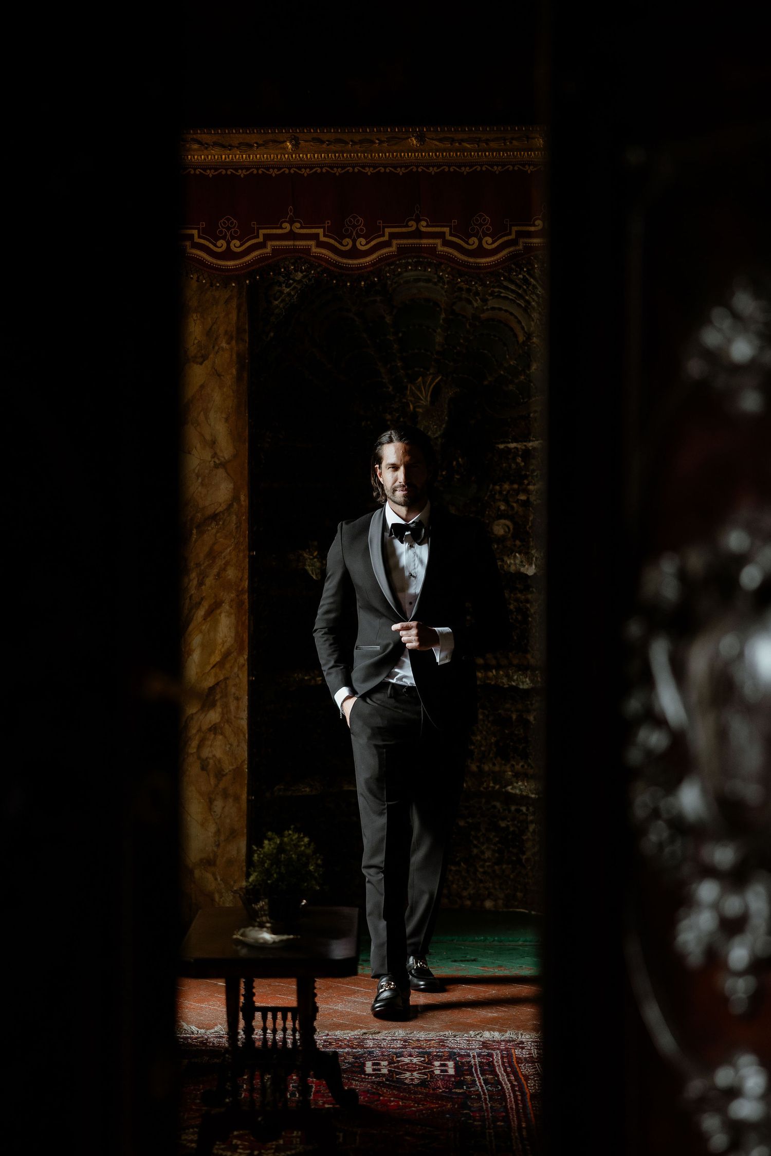 A man in a black tuxedo stands elegantly framed by an ornate doorway in dramatic lighting.