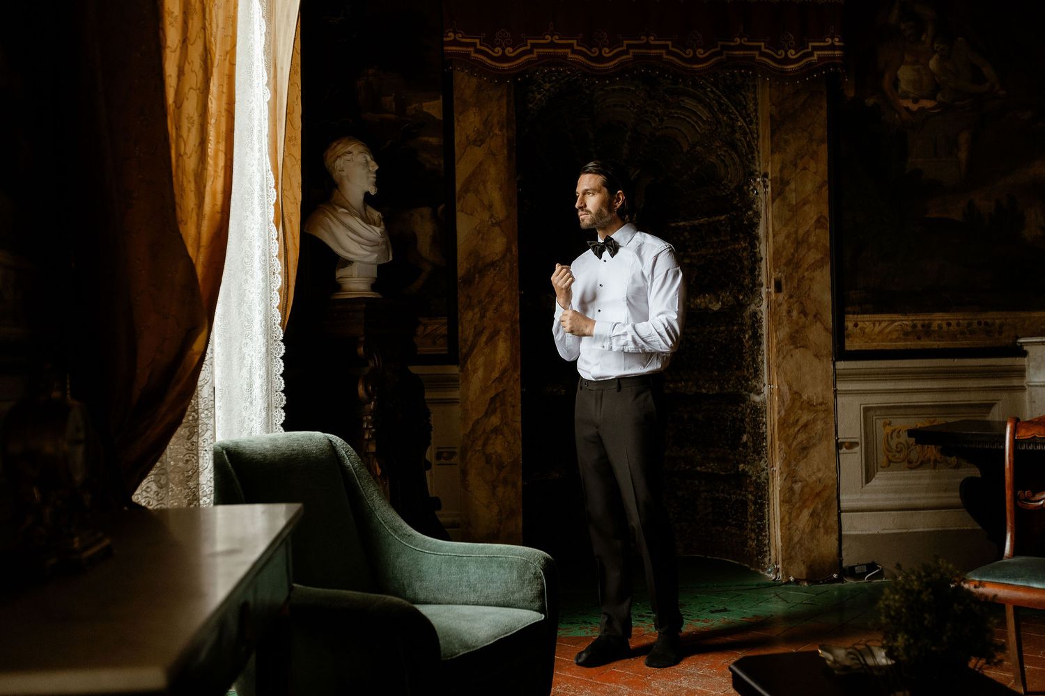 Someone in a white dress shirt and dark pants stands thoughtfully in an elegant vintage room with ornate decor and natural light.