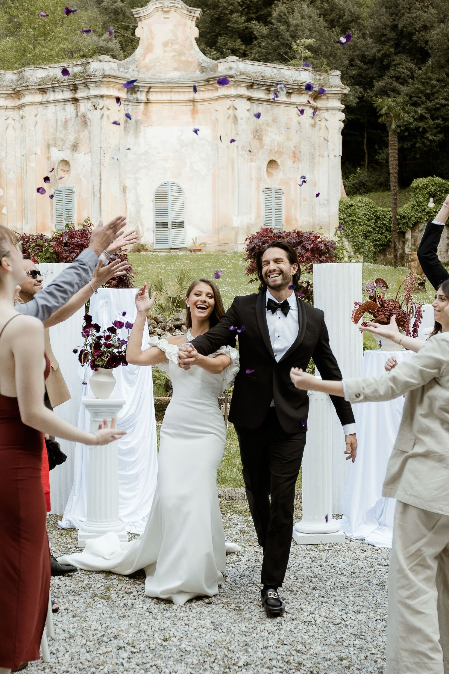 Wedding guests toss flower petals at a joyful couple exiting their ceremony in front of an ornate vintage building.