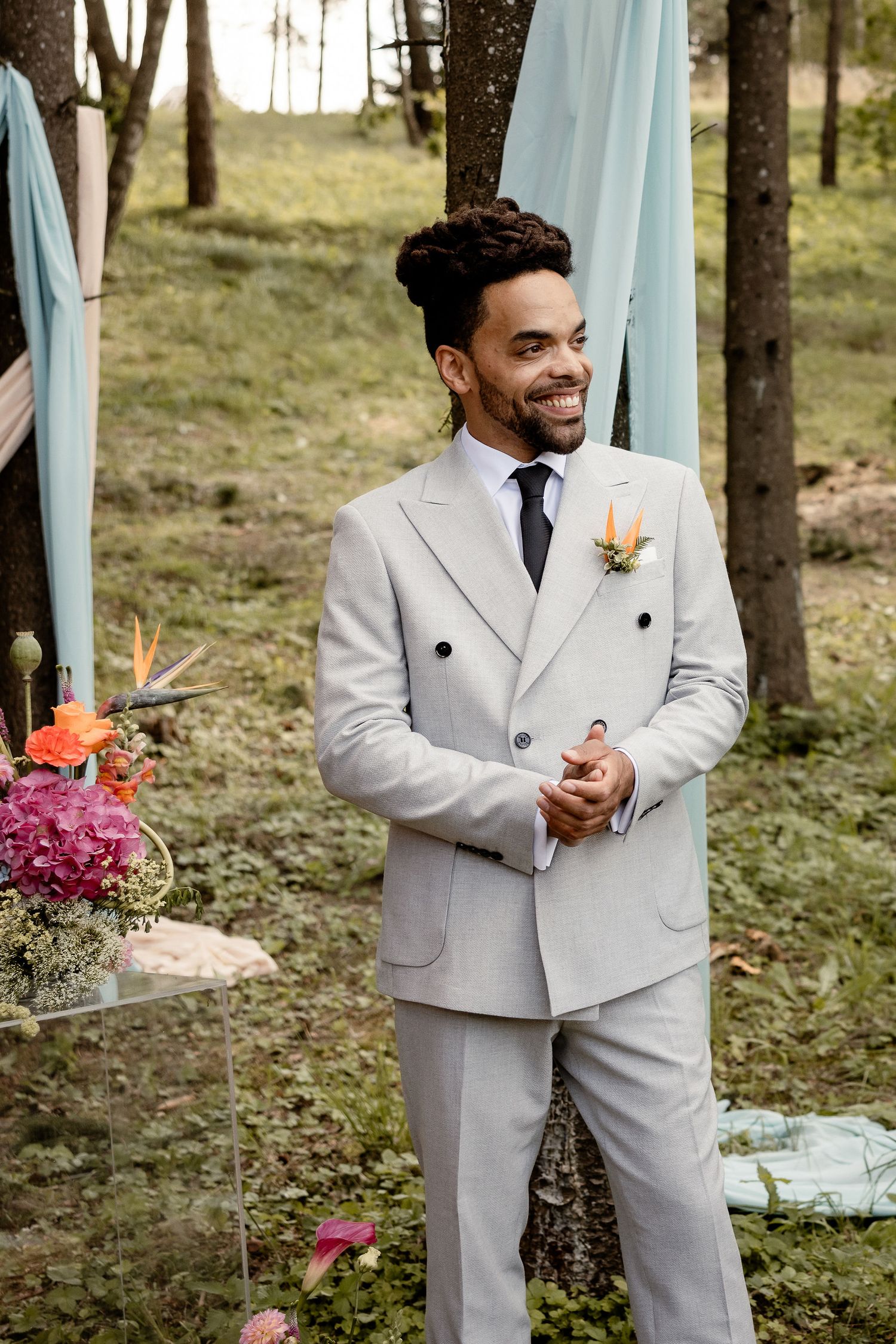 Groom in light gray double-breasted suit smiles during outdoor forest ceremony.