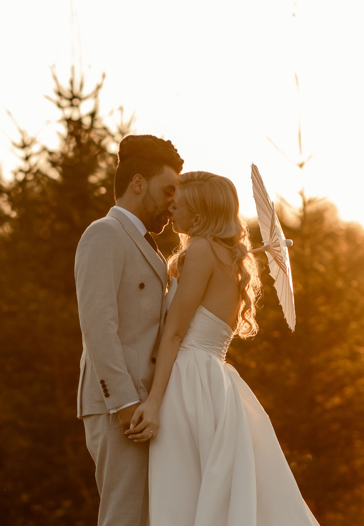 A romantic wedding couple shares an intimate moment at sunset with golden light filtering through the trees behind them.
