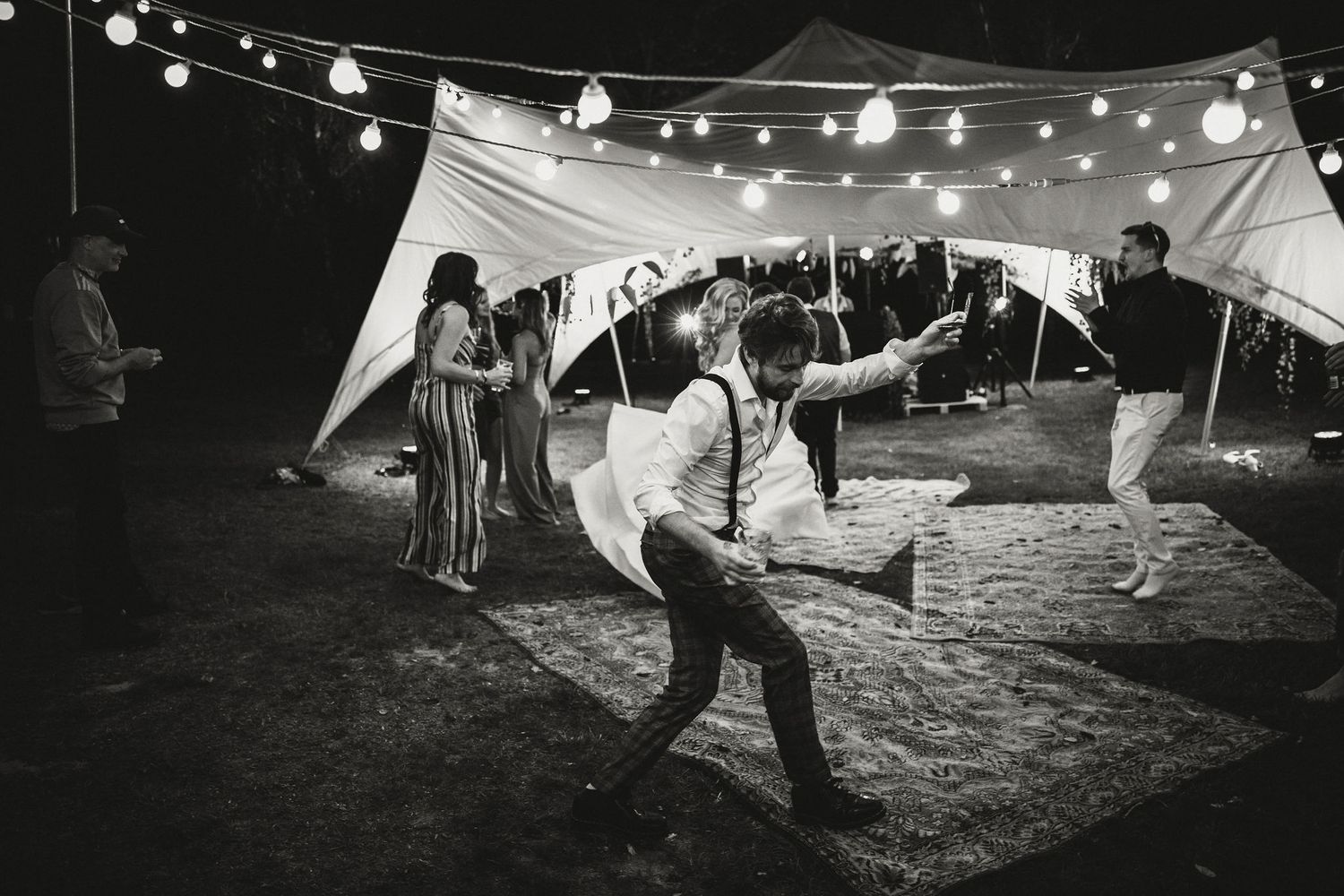 Dancers enjoying a lively celebration under a tent with string lights at an outdoor nighttime wedding reception.